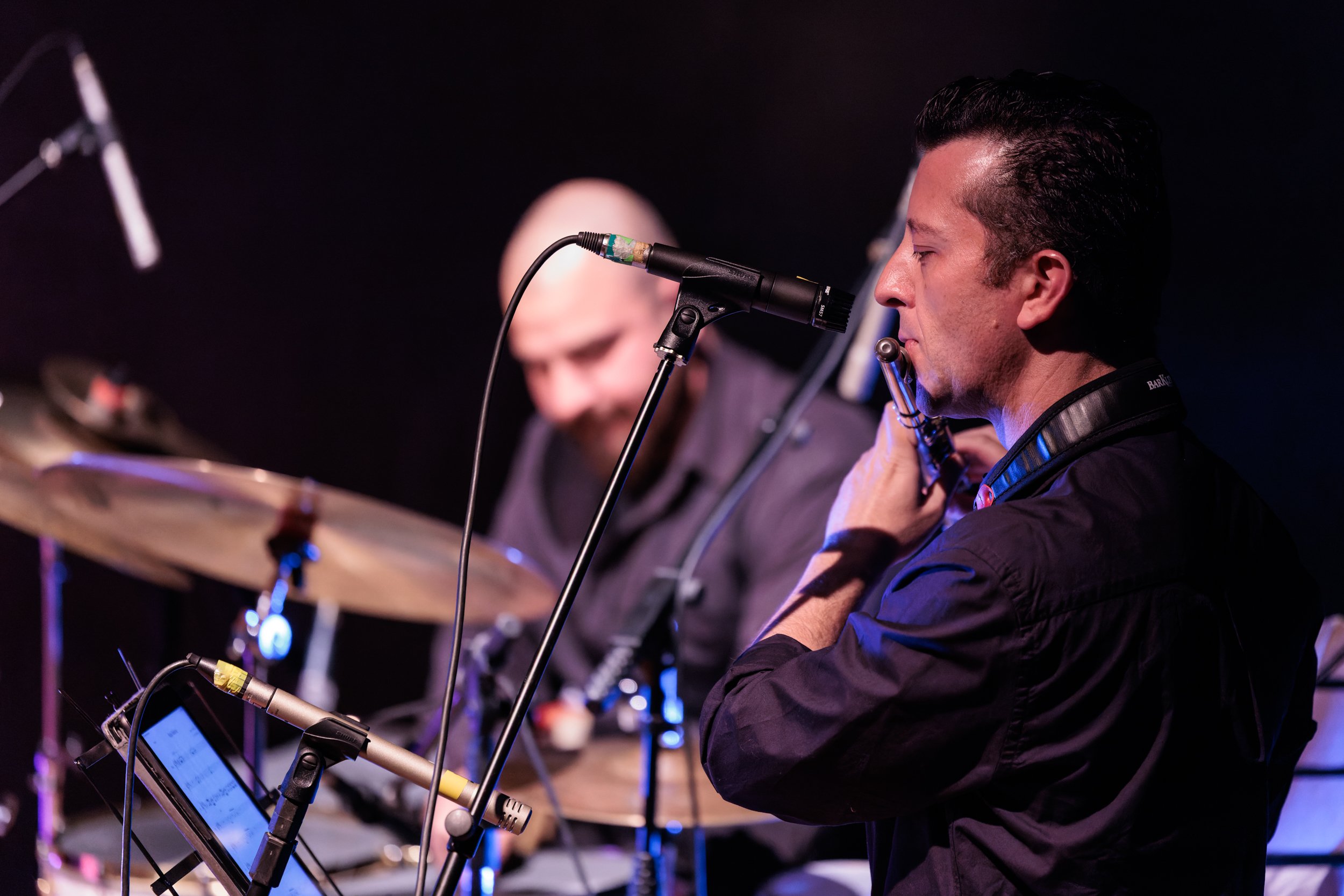 Musicians playing wind instruments during a live performance, with a microphone and electronic device in front of them.
