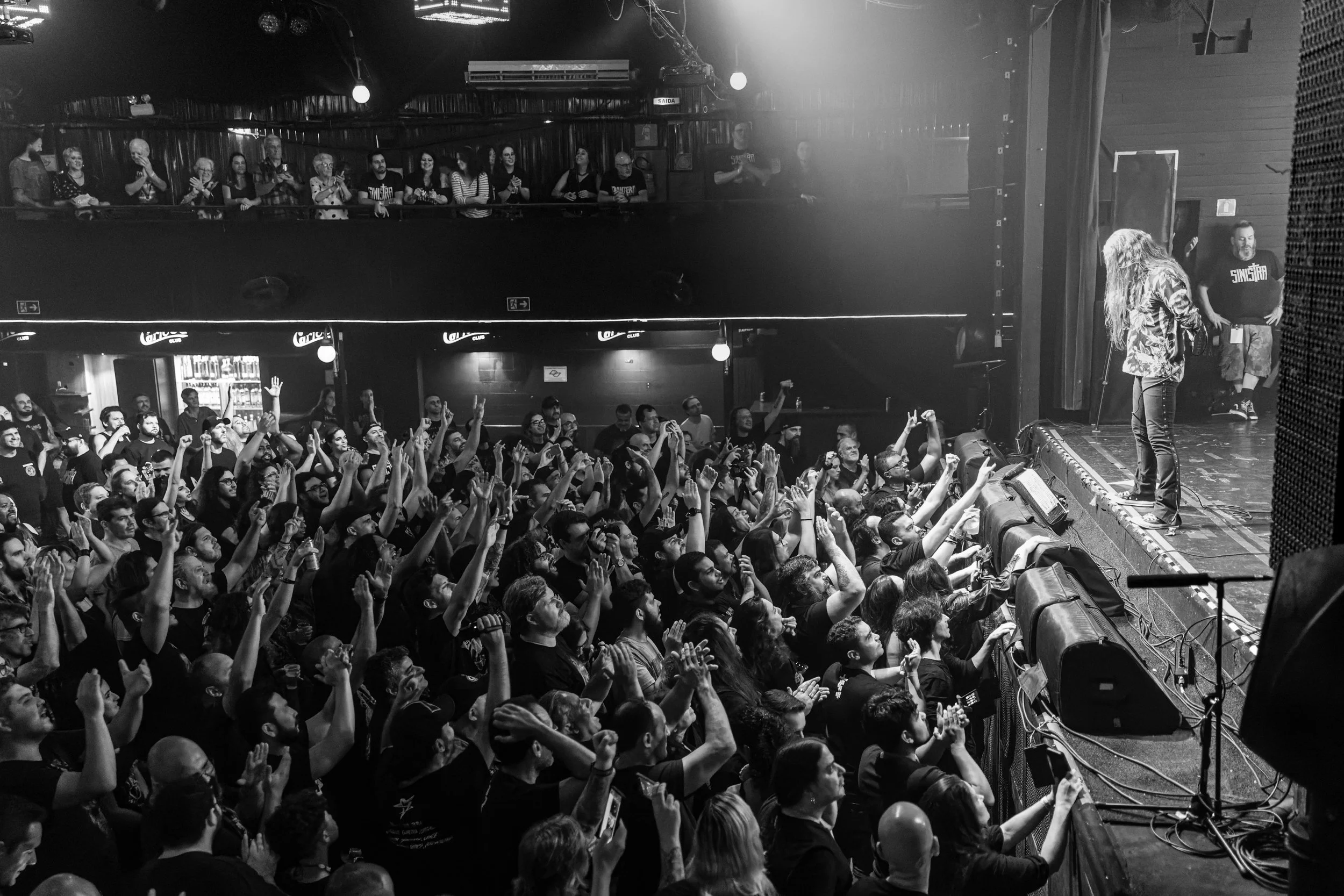 A concert with a large audience in front of a stage. The audience members are clapping and raising their hands. A performer is standing on stage near a microphone.