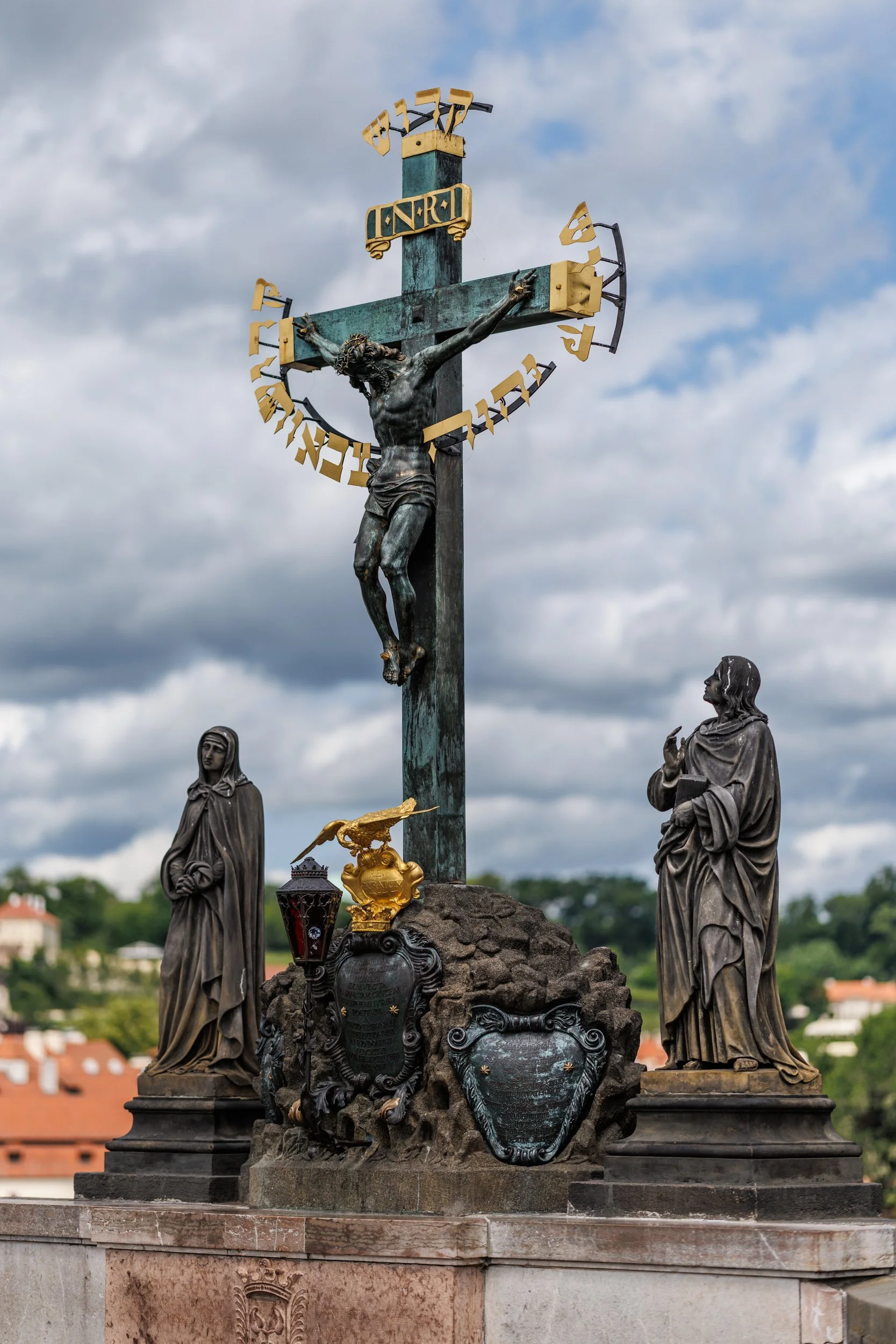 A religious crucifix monument with a sculpture of Jesus on the cross, surrounded by statues of religious figures, against a cloudy sky background.