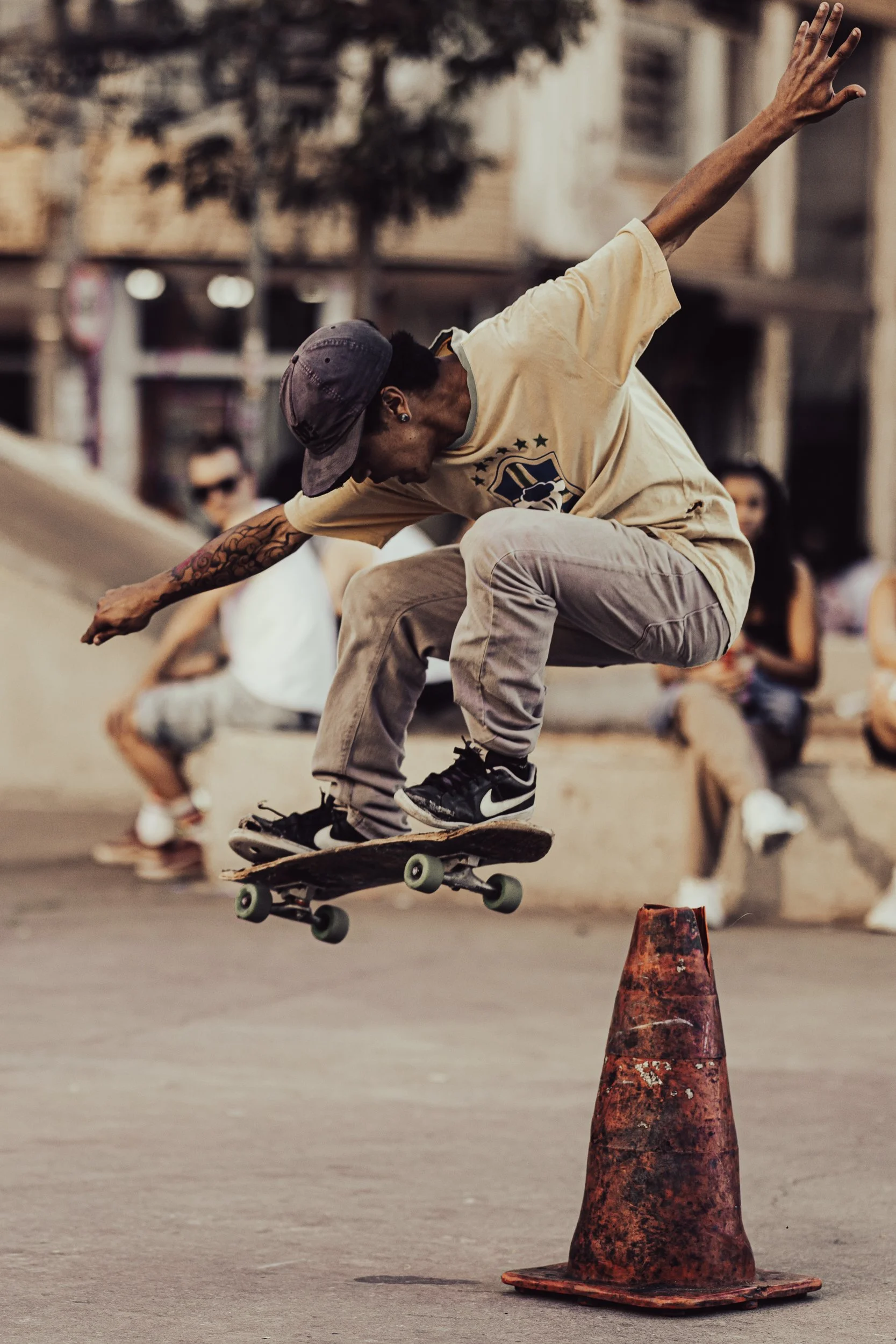 A skateboarder catches air while jumping over an orange traffic cone on a city street, with blurred spectators in the background.