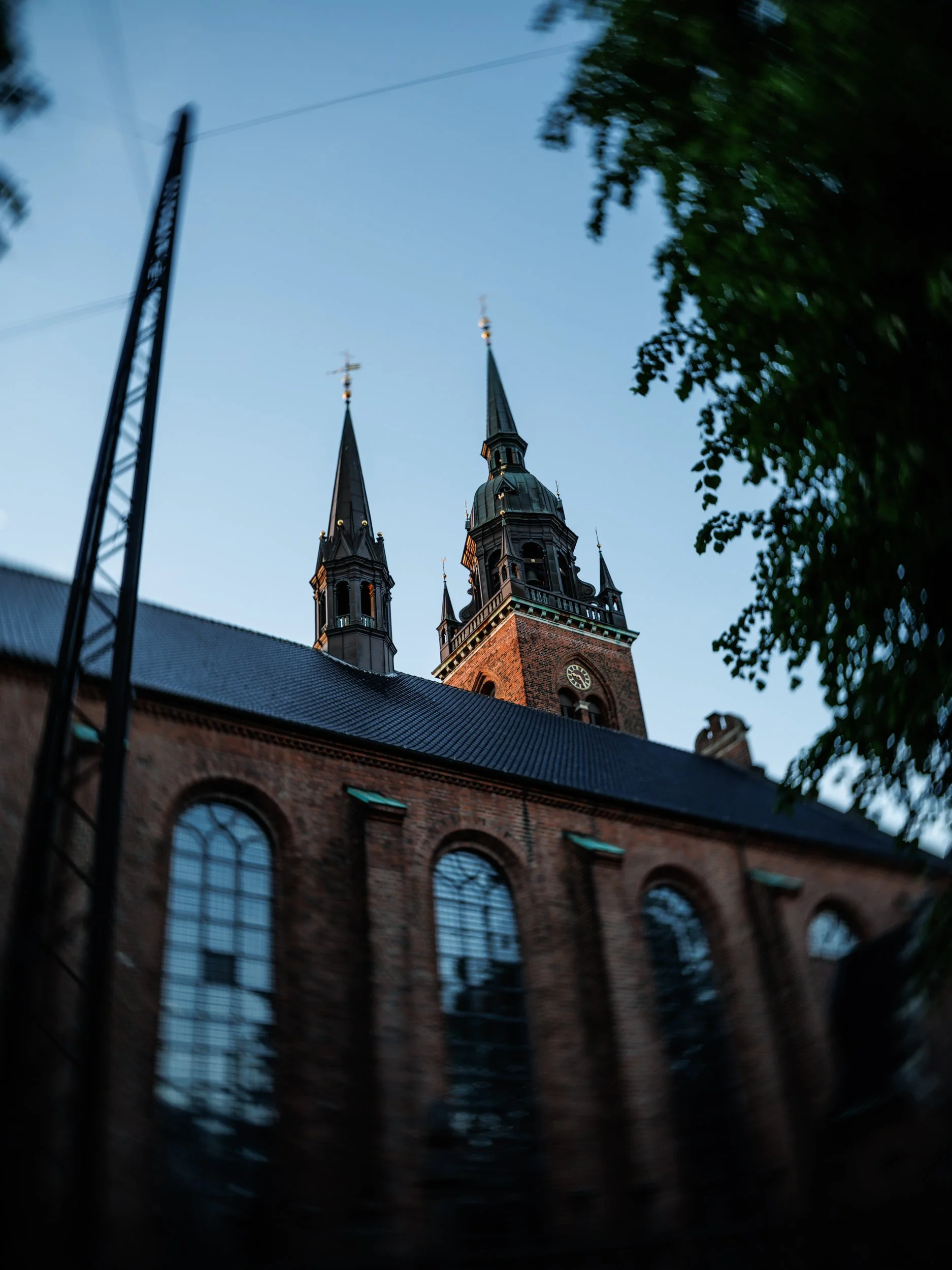 A church with tall steeples viewed from below, with a blue sky and some trees framing the scene.