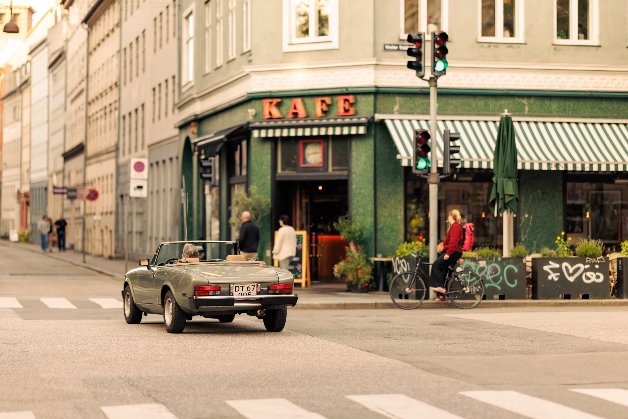 A vintage convertible car driving down a city street with a restaurant on the corner that has a green exterior and red sign reading 'KAFFE'. Pedestrians are walking and standing near the restaurant, and there are bicycles parked nearby.