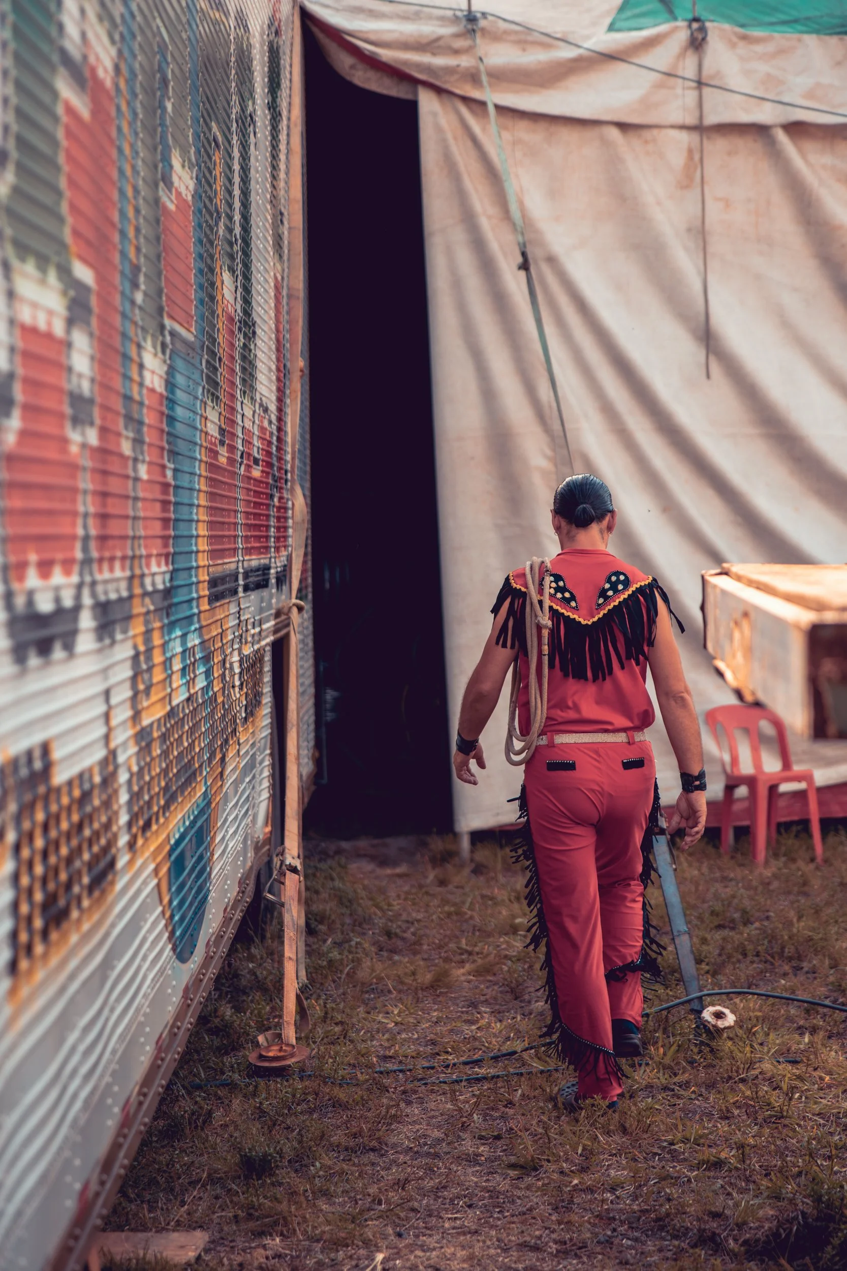 A woman dressed in red with black fringe details walks away from the camera past a large trailer with colorful patterns on its side, towards an open tent or structure. She has her hair tied in a bun and is carrying some ropes.