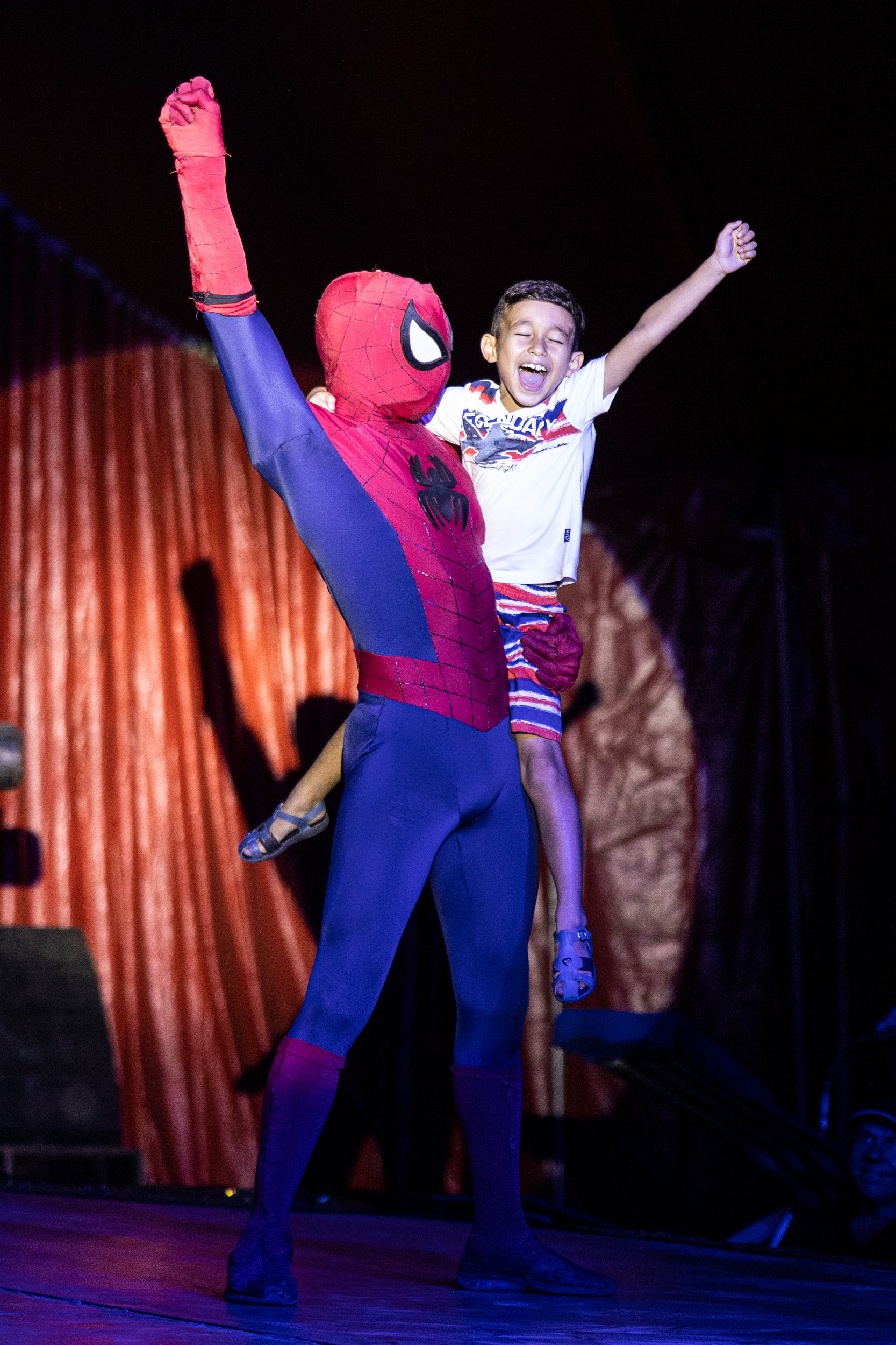 A person dressed as Spider-Man lifting a young girl with raised arms, both appearing joyful, on a stage with red curtains in the background.