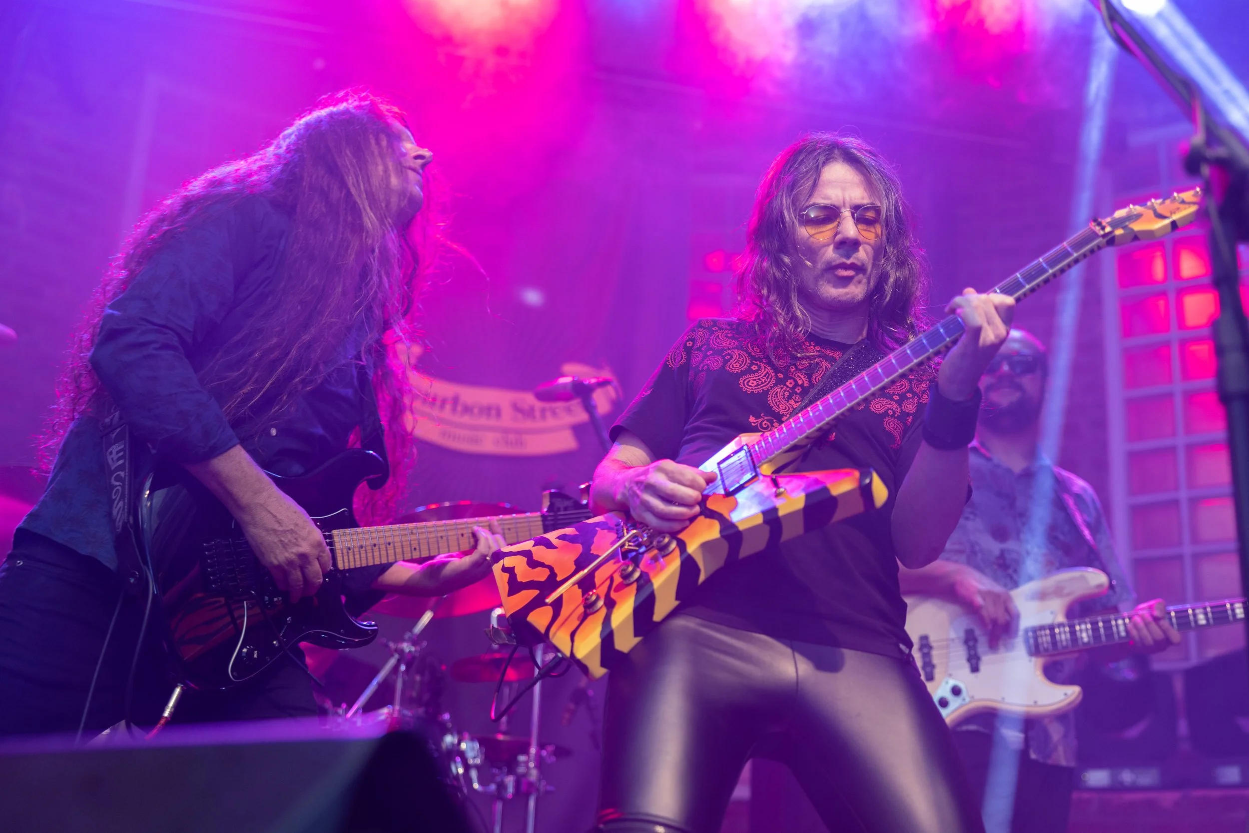 Two musicians performing on stage with colorful lighting. One is playing an electric guitar, the other a keytar, with a background of other band members and stage equipment.