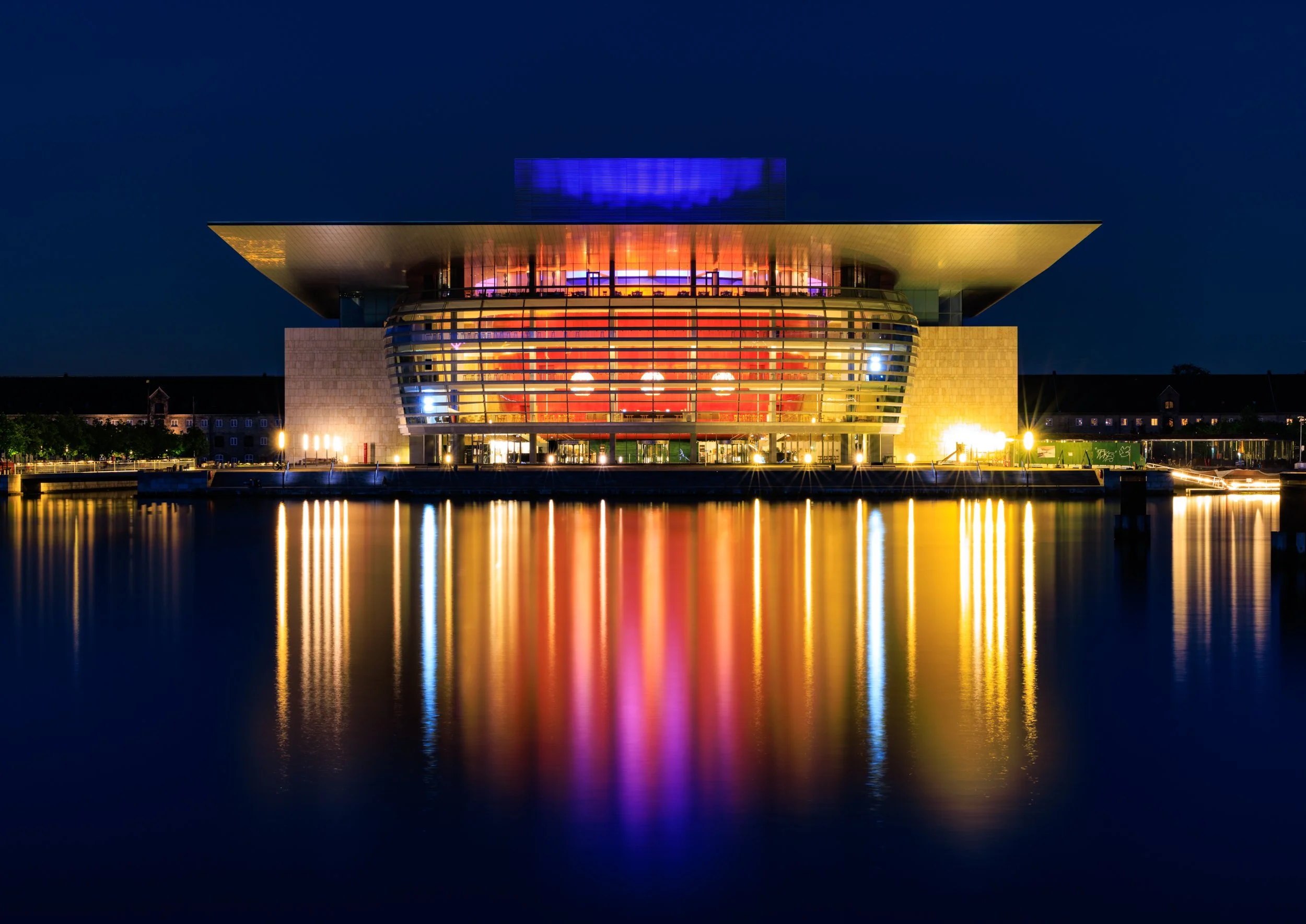 Night view of a modern building with colorful lights reflecting on the water in front of it.