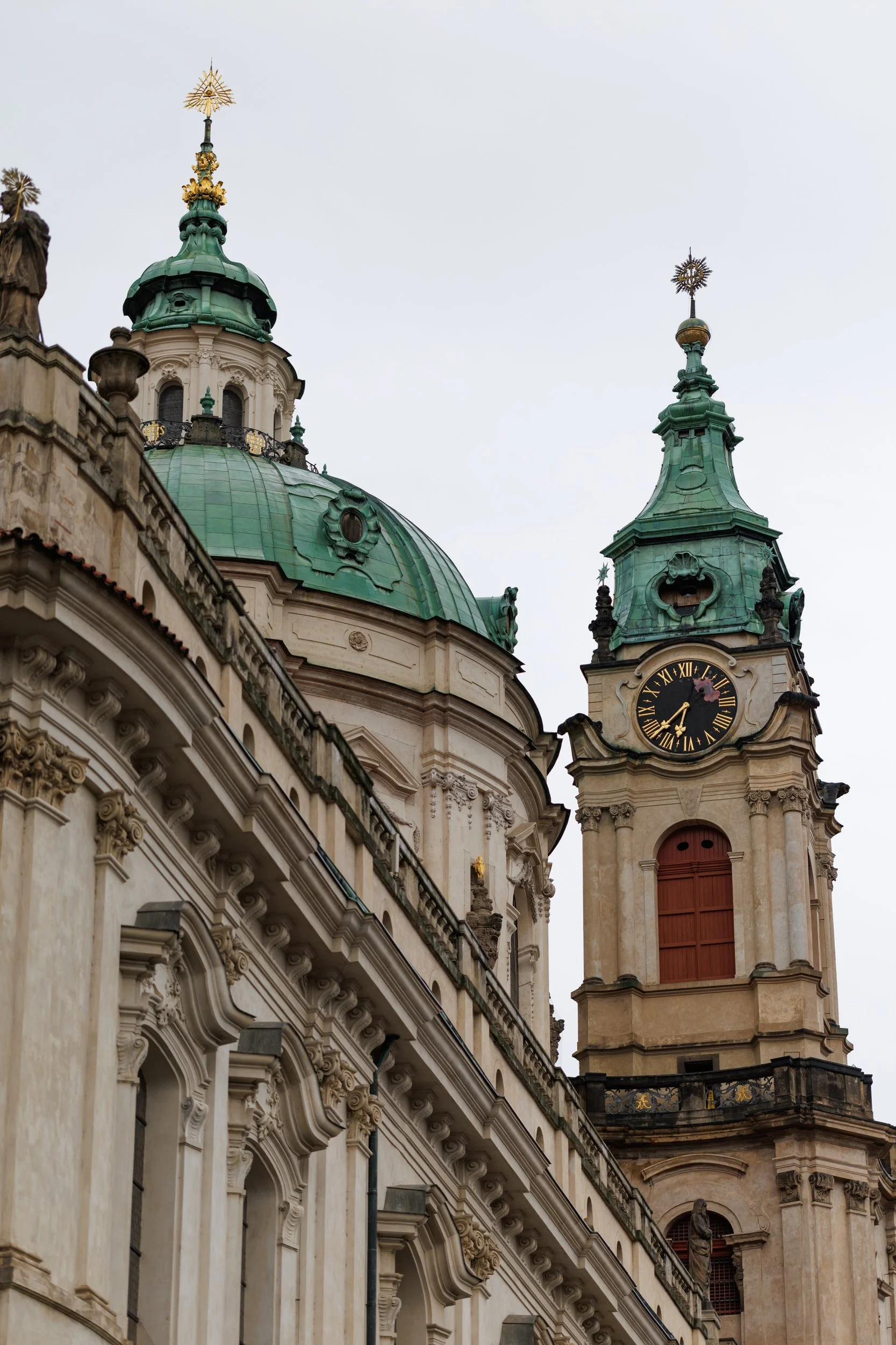 Close-up of an ornate historic church with green domed roofs and clock tower, featuring statues and decorative architectural details, under a cloudy sky.