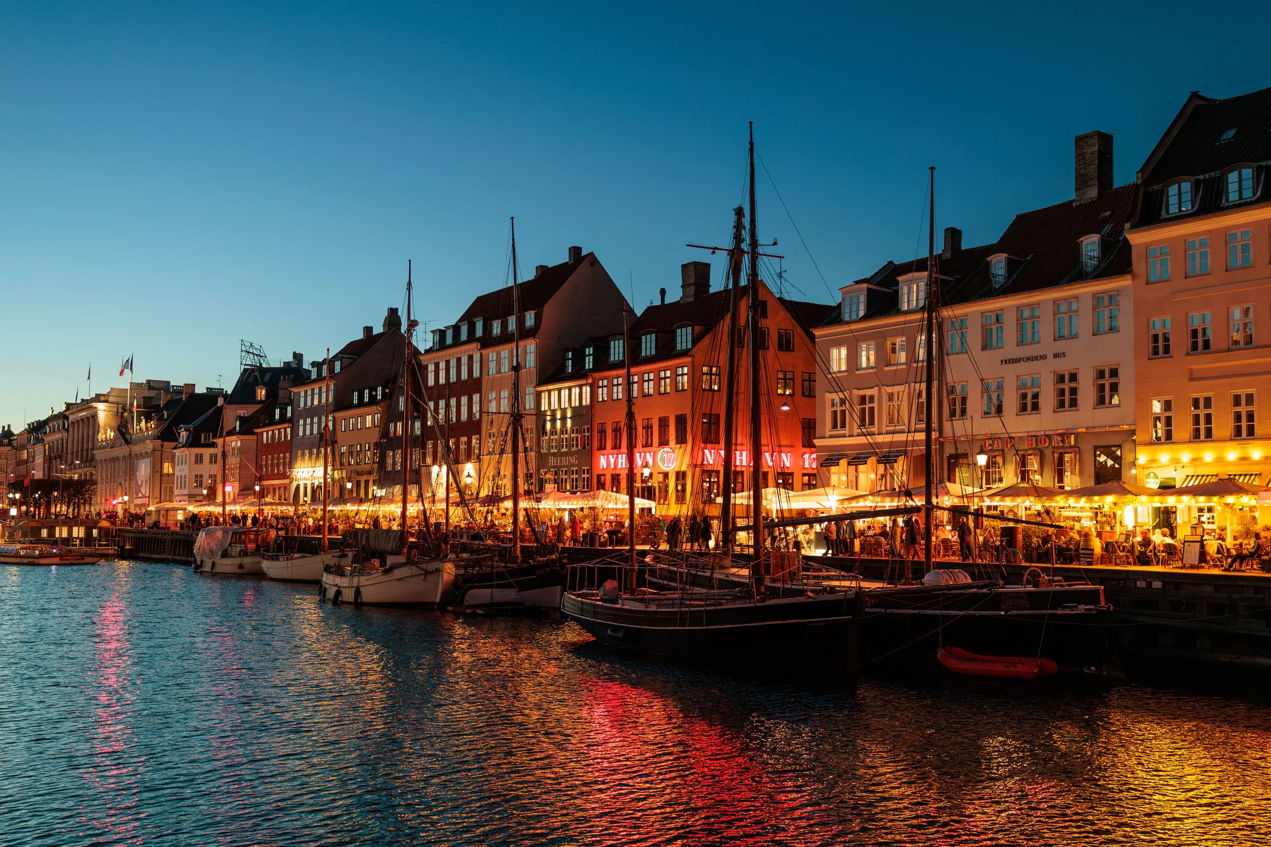 A waterfront view of a cityscape at dusk, with colorful buildings, lit signs, and boats docked on the water.