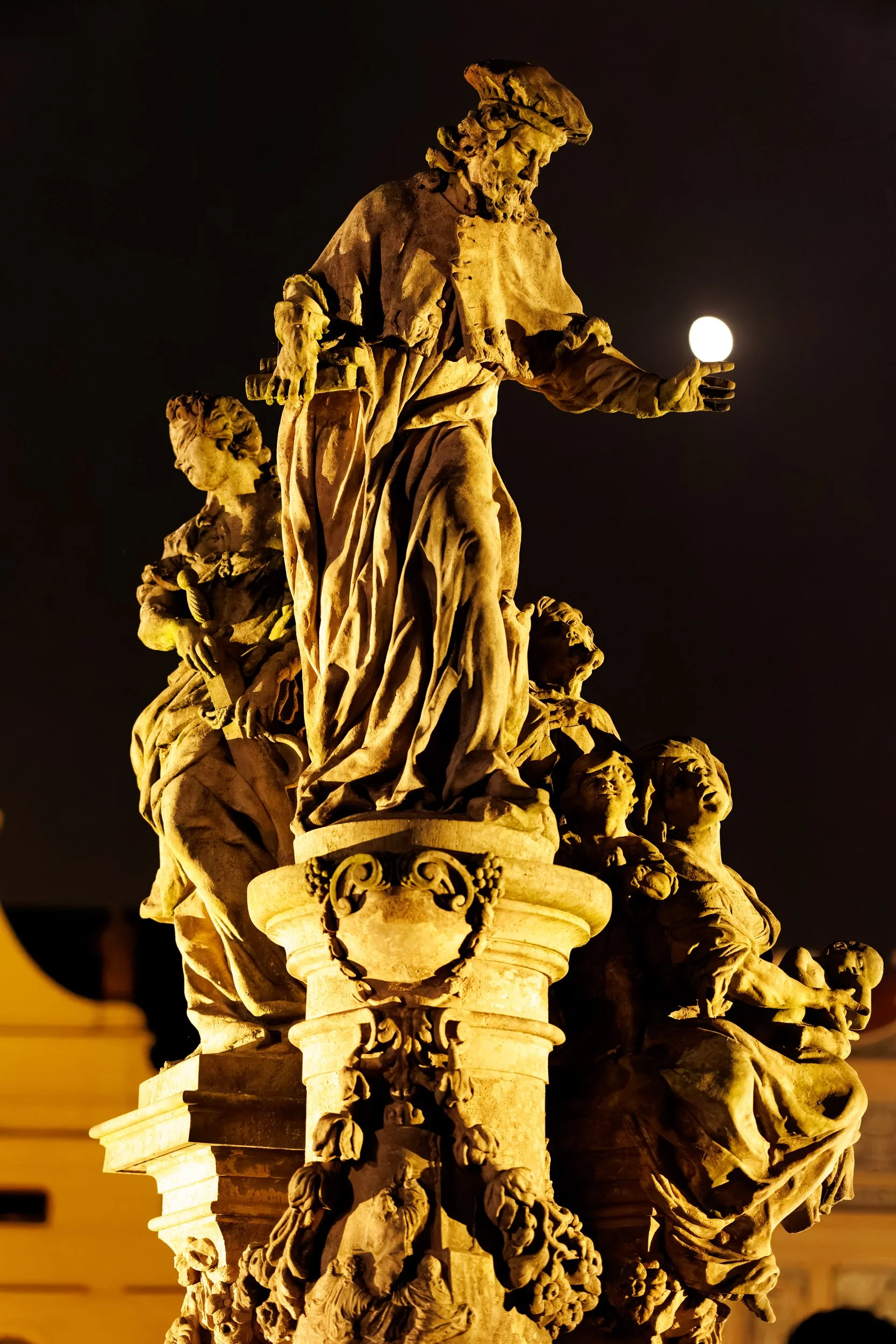 Nighttime view of a detailed stone statue depicting a person holding a sphere, surrounded by other figures and ornate decorative elements.