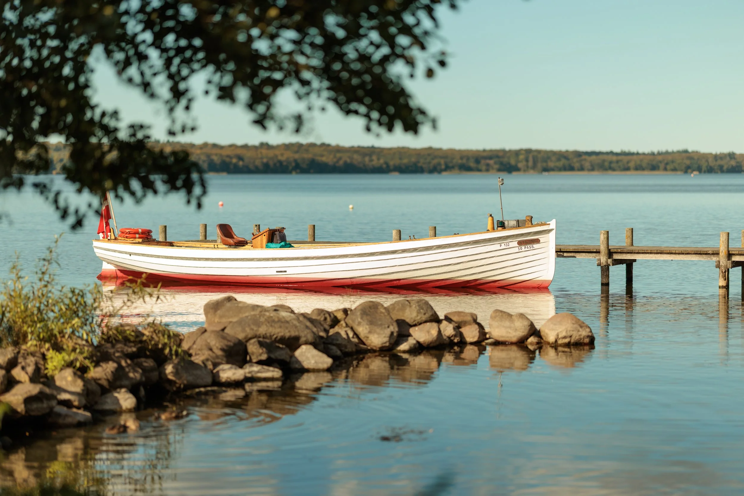 A white boat with a red bottom docked at a wooden pier on a calm lake, framed by rocks and trees in the foreground with a distant shoreline in the background.