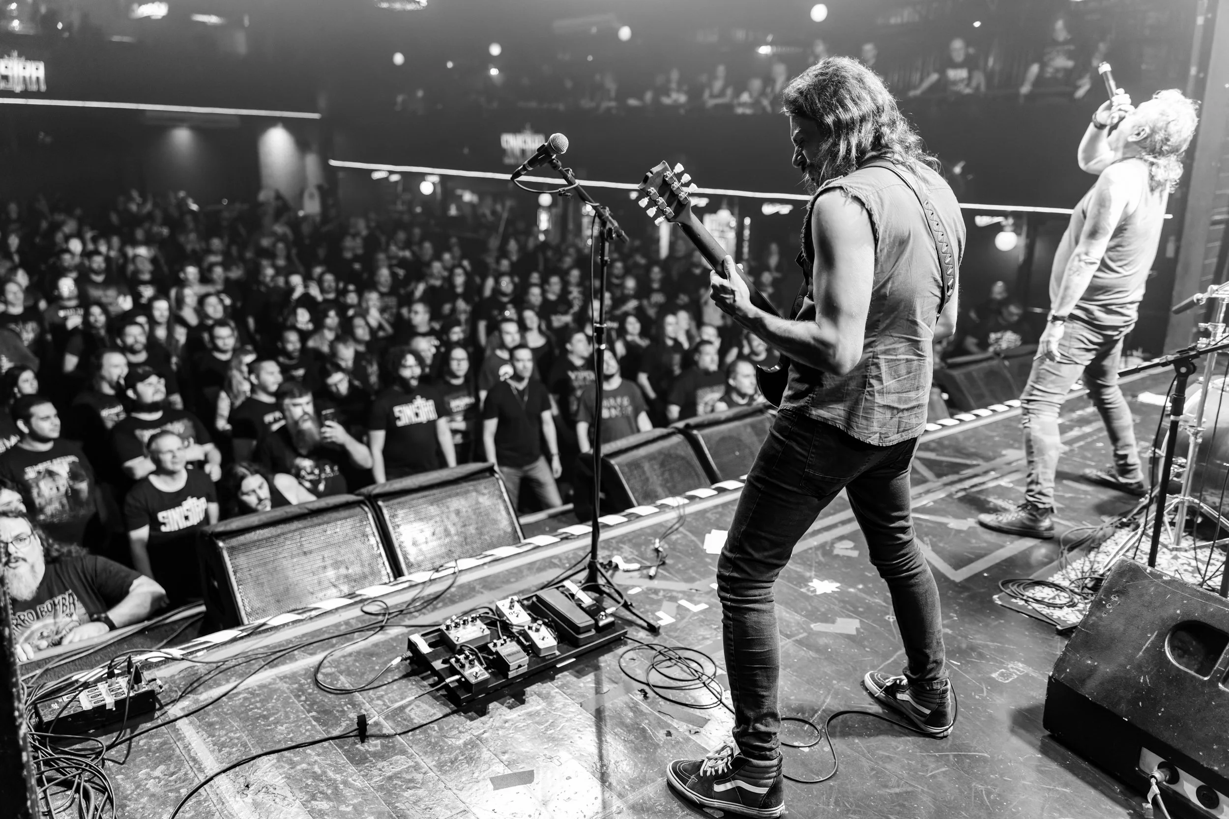 Black and white photo of musicians performing on stage in front of a large audience. One musician is playing an electric guitar, another is singing into a microphone, and a third is playing drums. The crowd is watching attentively inside a concert ve