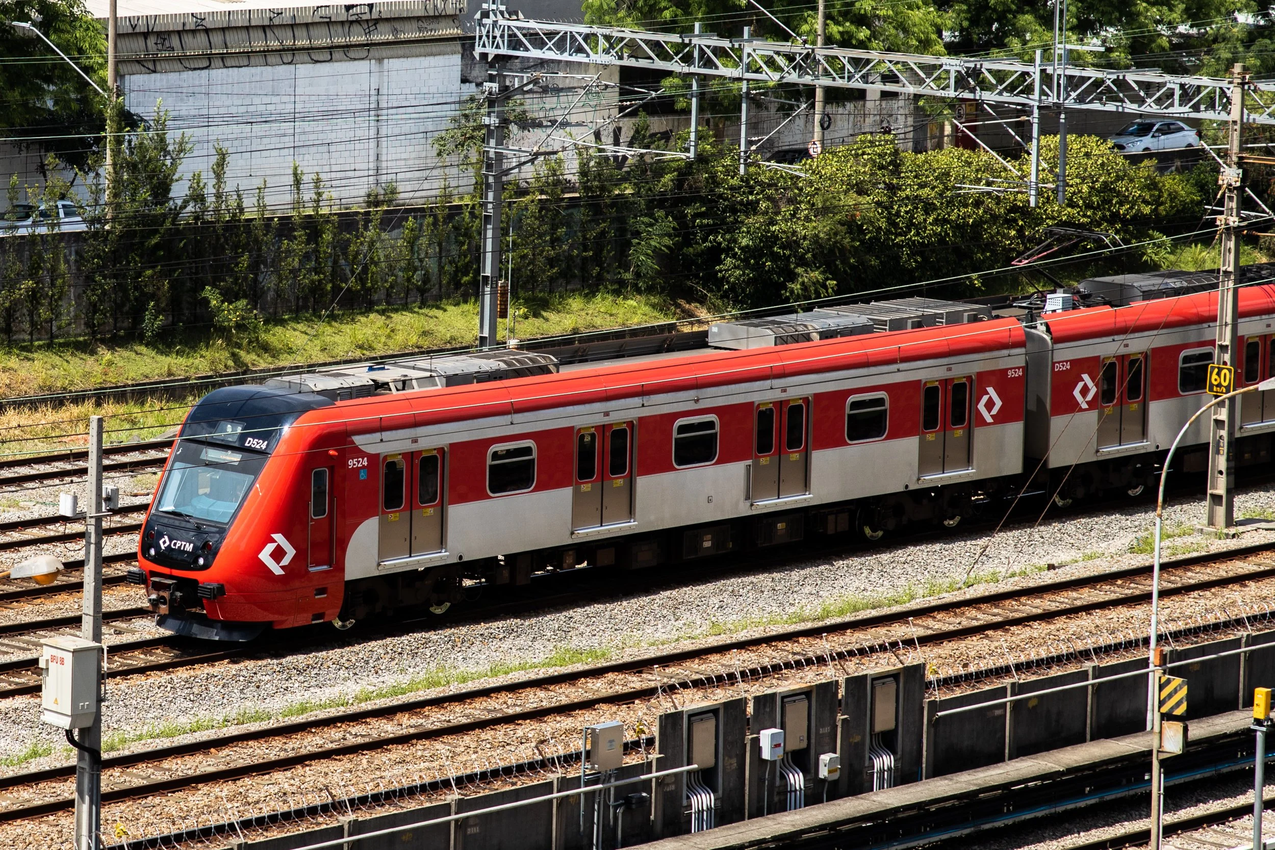 A red train traveling on railway tracks, overhead electrical wires, green trees, and cars parked behind a fence in the background.