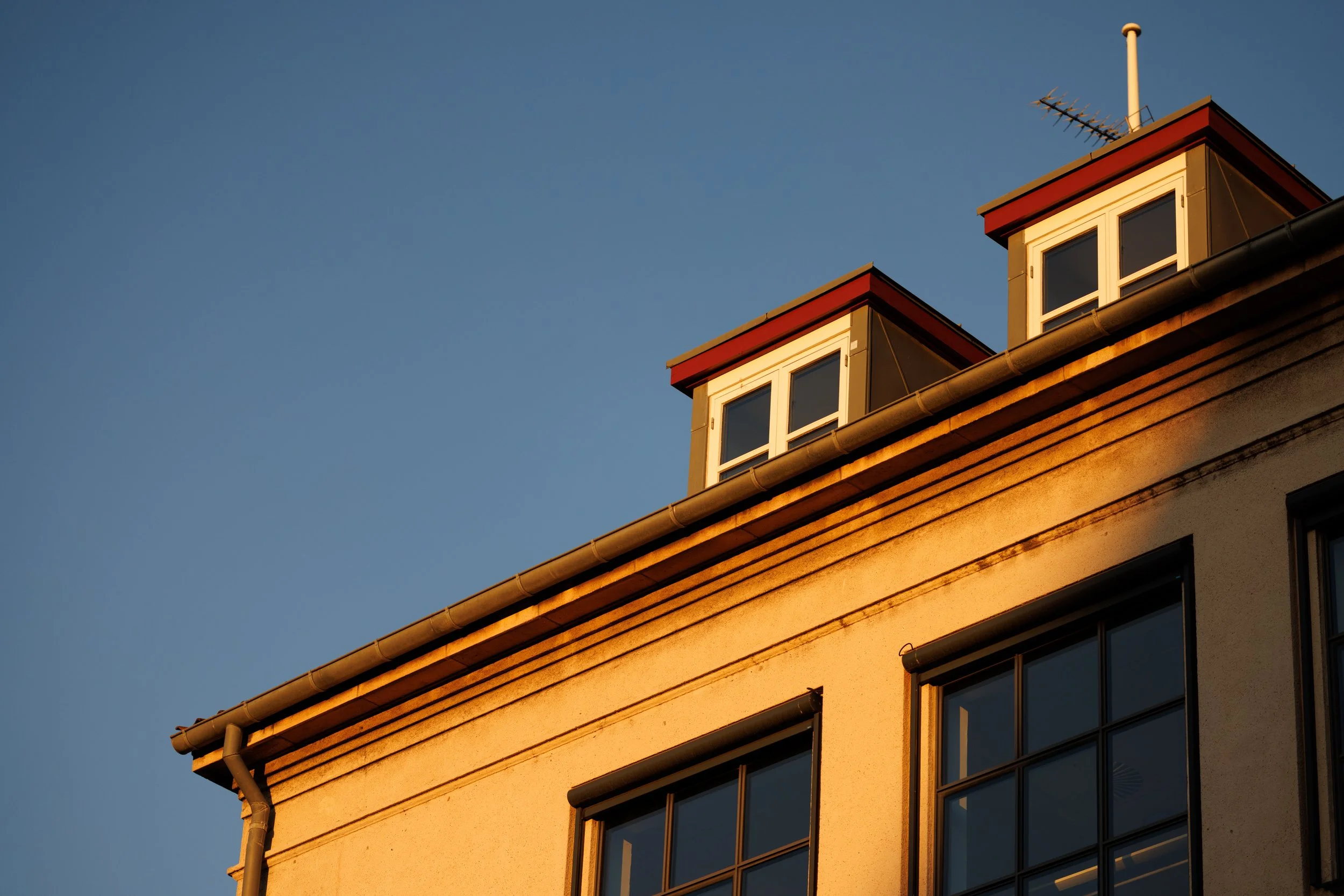 Close-up view of a building's corner with windows, tan walls, a red trim at the roof, and a satellite antenna on top, bathed in warm sunlight against a clear blue sky.