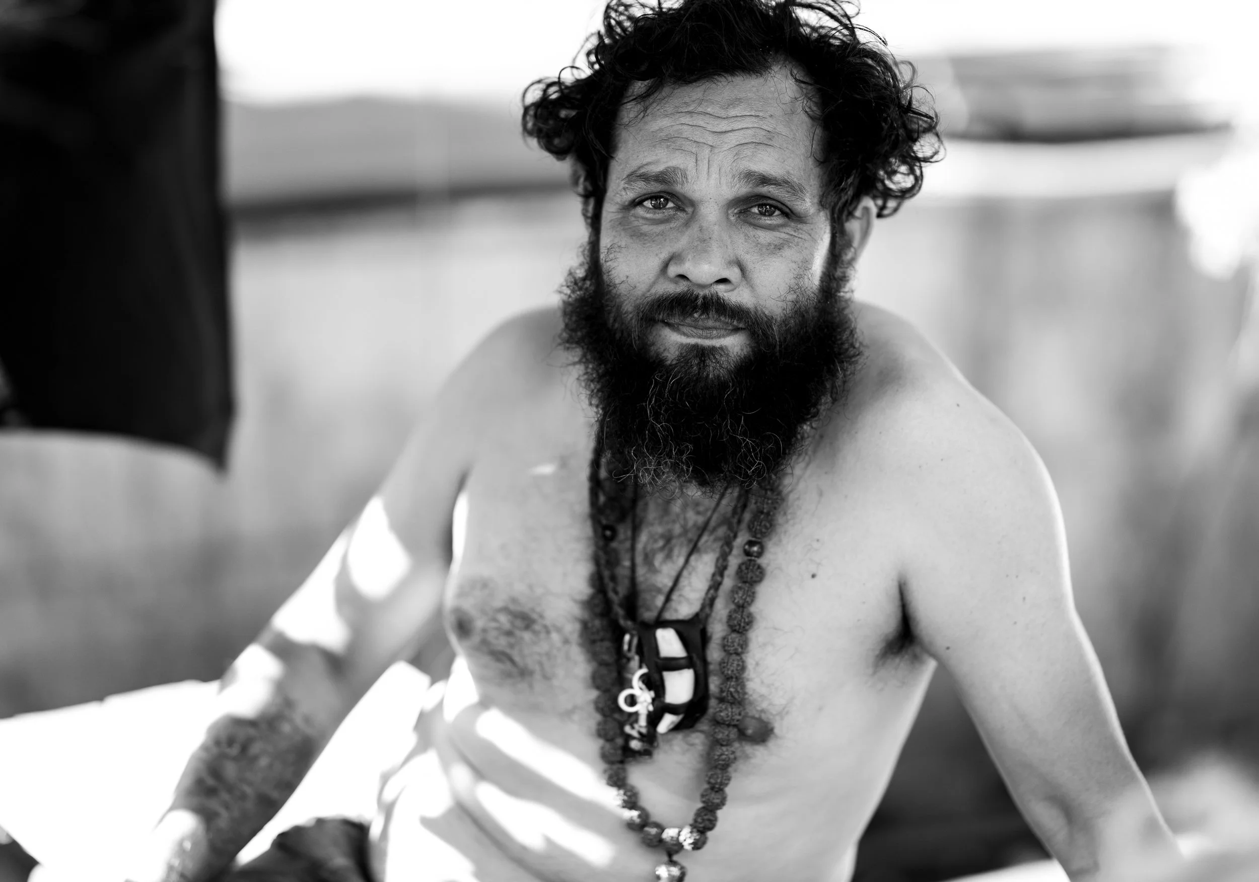 A black and white photo of a shirtless man with a beard and curly hair, wearing multiple necklaces, looking at the camera with a slight smile.