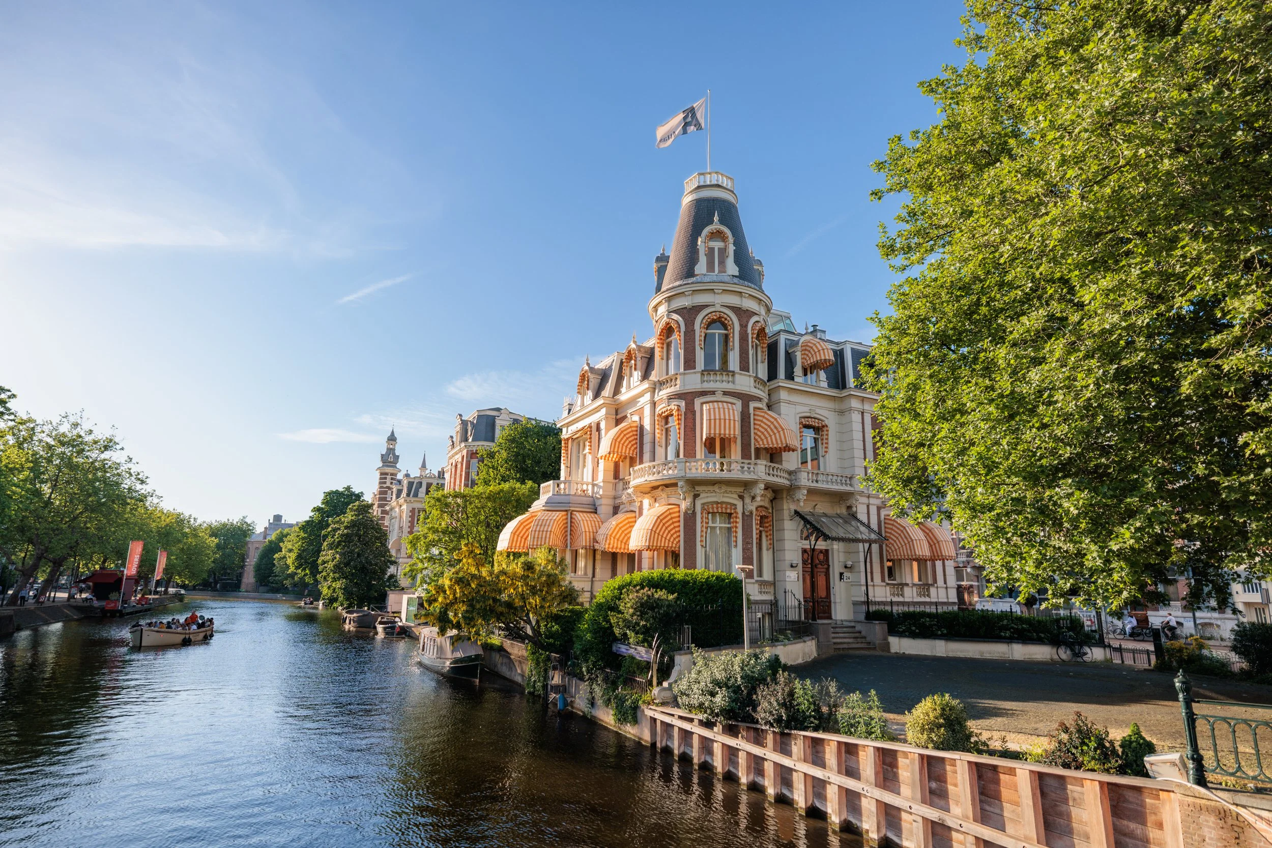 A historic European-style building with a turret and striped awnings along a canal, surrounded by green trees and a clear blue sky, with boats on the water