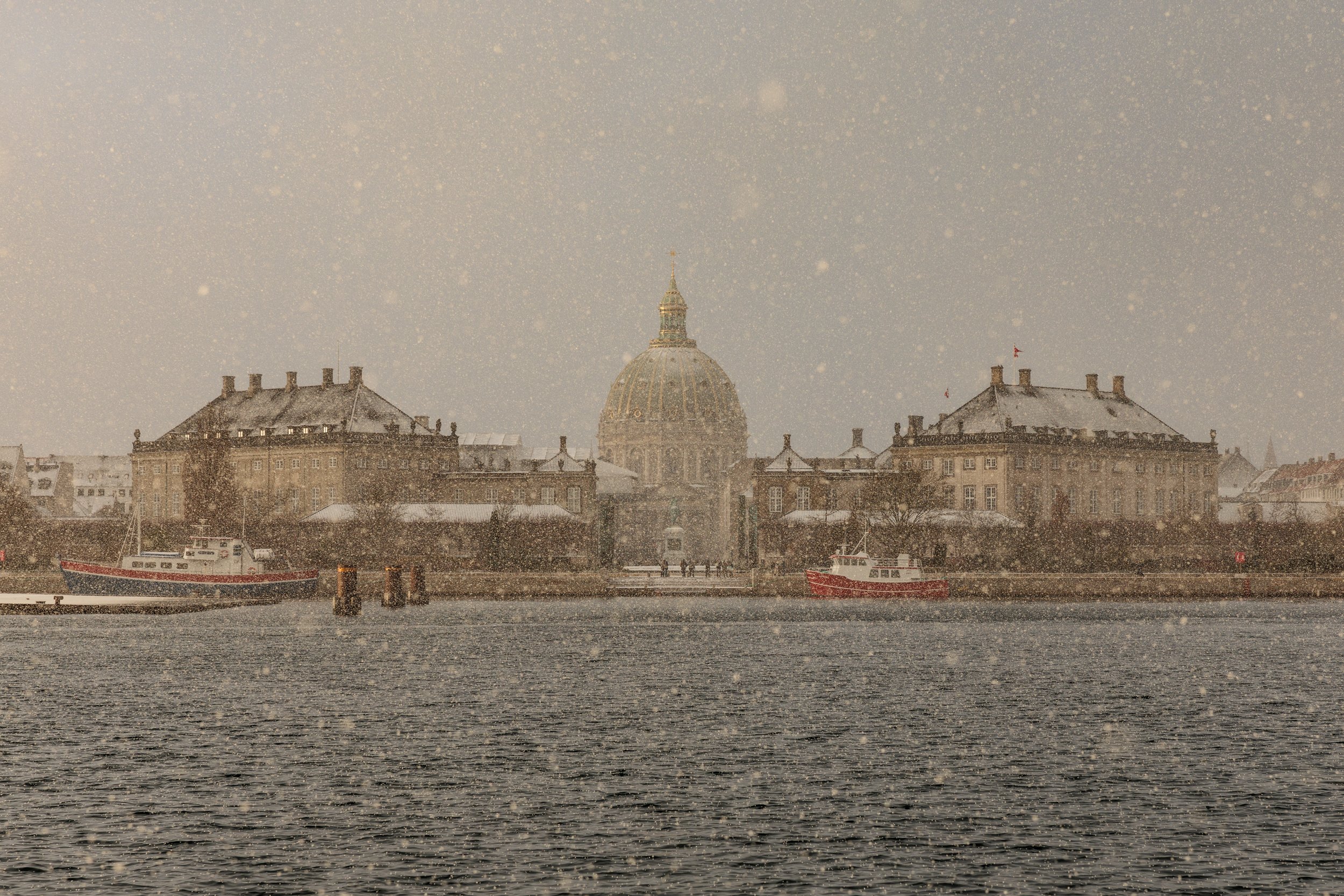 Snow falling over historic buildings and boats docked along a waterfront, with prominent dome and flags in the background.