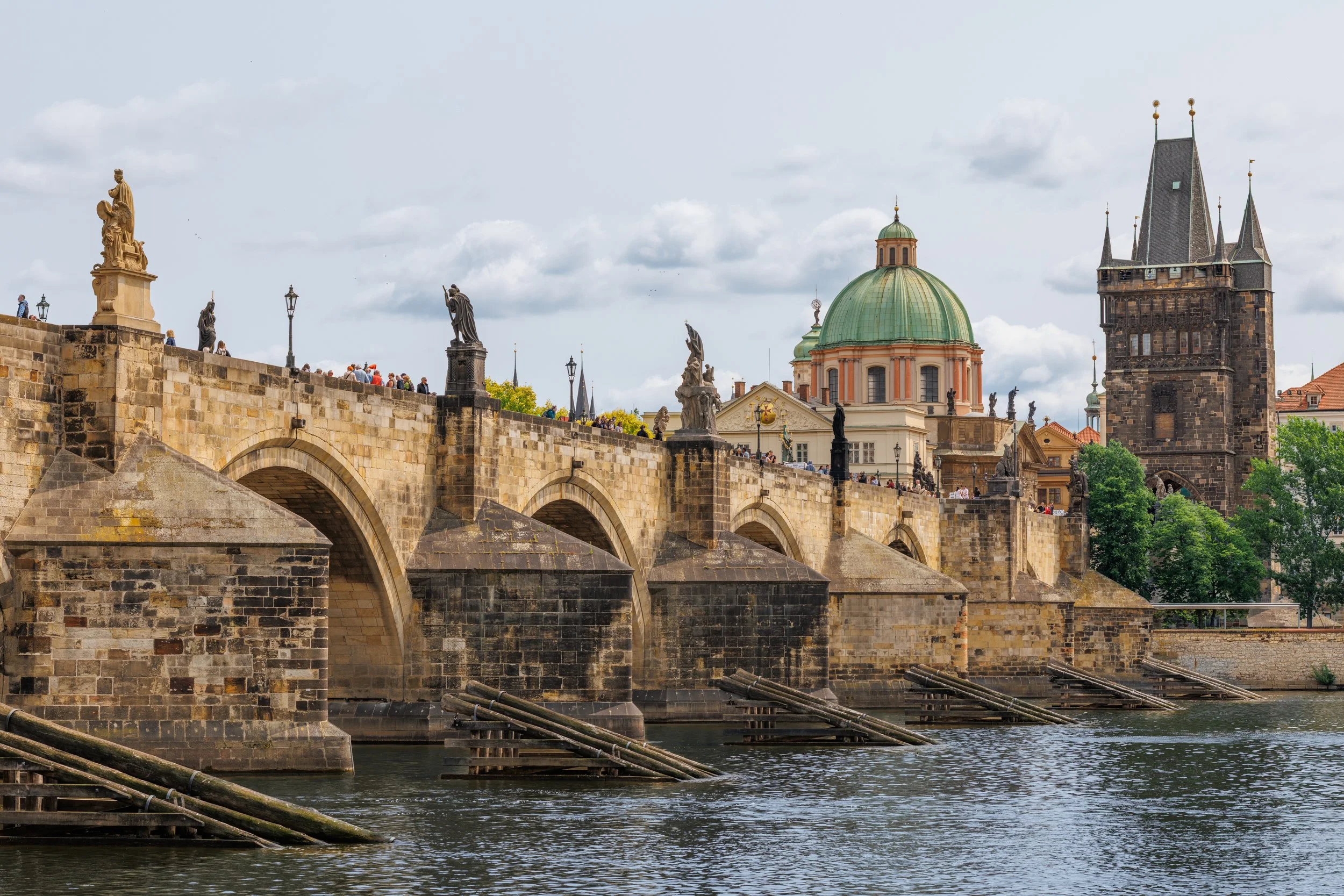 View of Charles Bridge in Prague, Czech Republic, with stone arches over the Vltava River, adorned with historical statues, and a cathedral with a green dome and tall spires in the background.