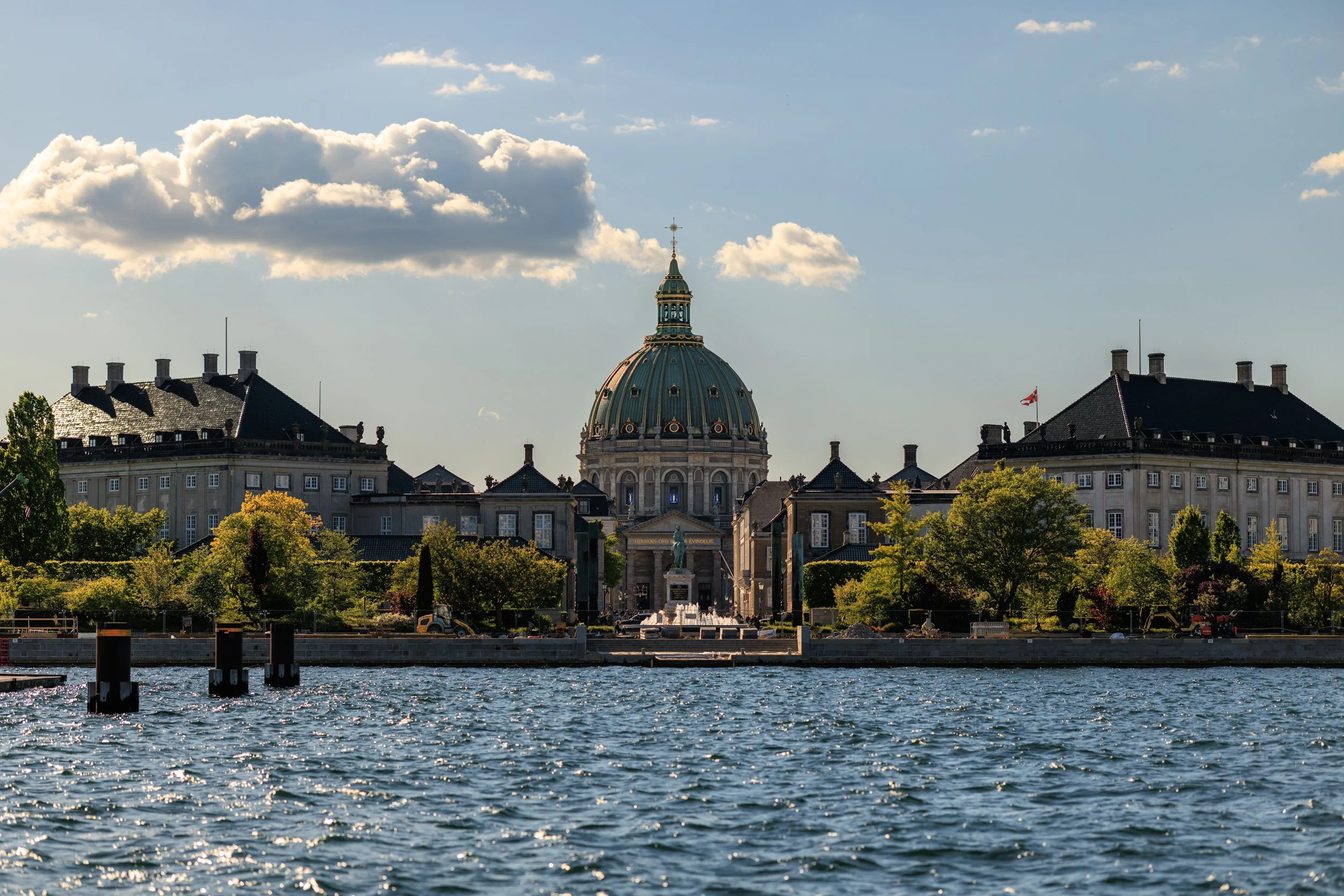 View of a prominent domed historic building with a statue at the entrance, surrounded by trees, as seen across a body of water with sunlight reflecting on the surface, under a partly cloudy sky.