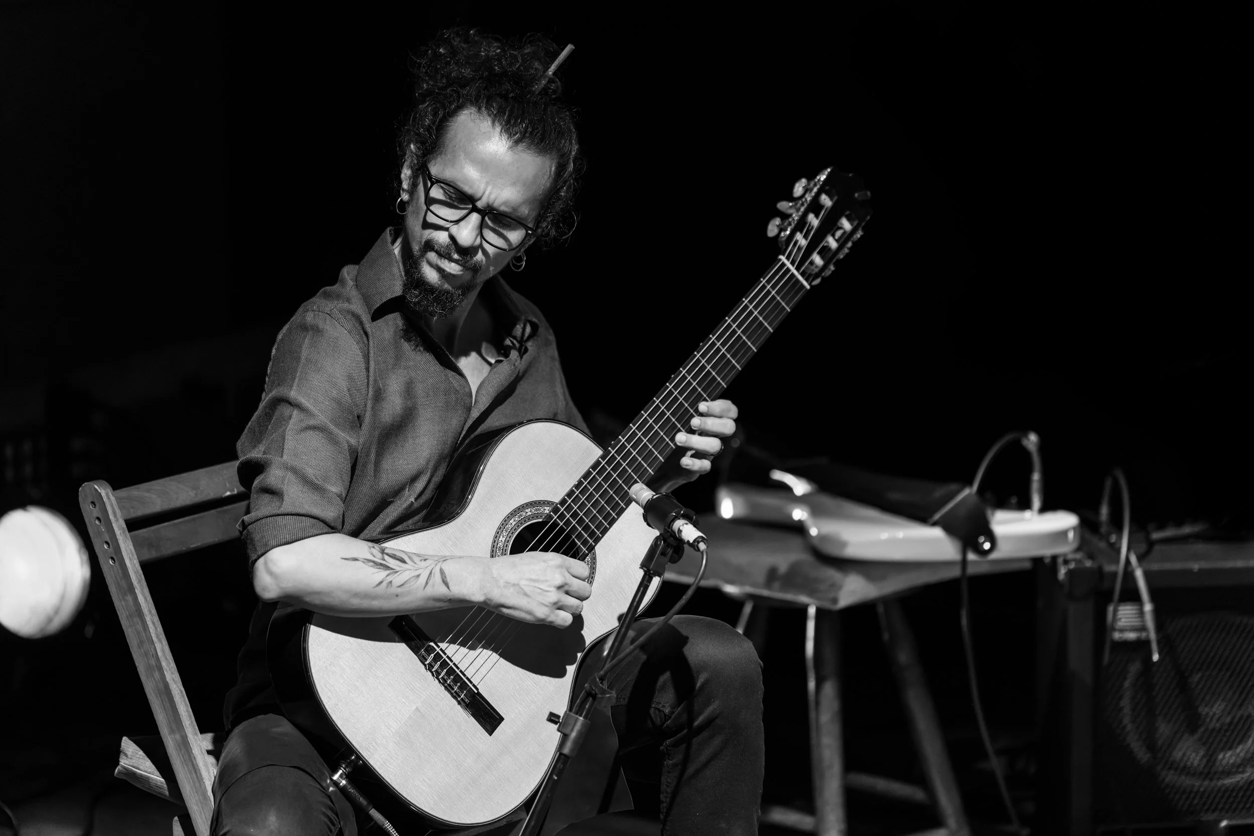 A man with curly hair, glasses, and a beard plays an acoustic guitar on stage. He is seated on a wooden chair, with a microphone in front of him. The background is dark, with some electronic equipment on a table nearby.