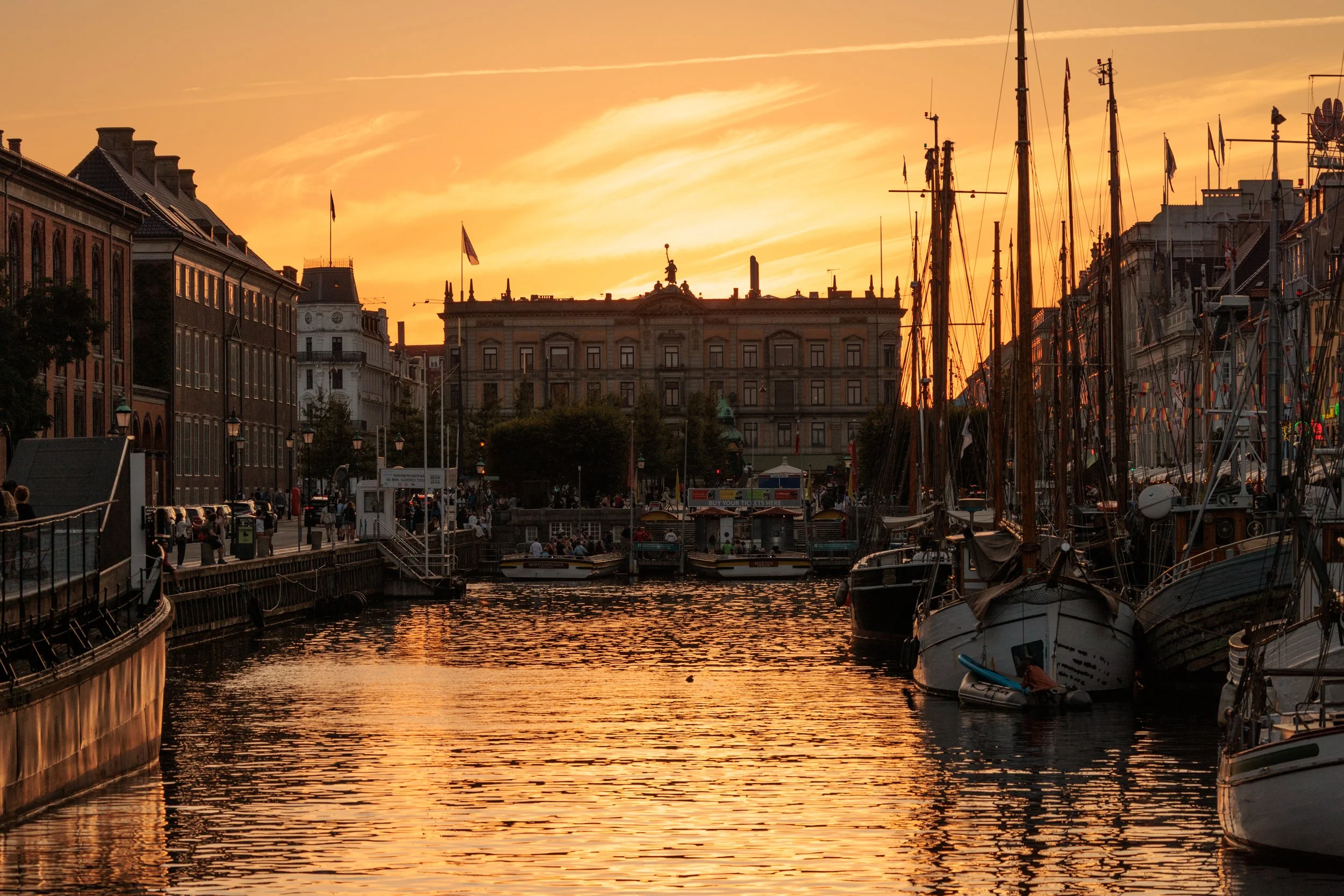A harbor scene at sunset with sailboats docked, historic buildings, and a sunset sky with orange and yellow hues.
