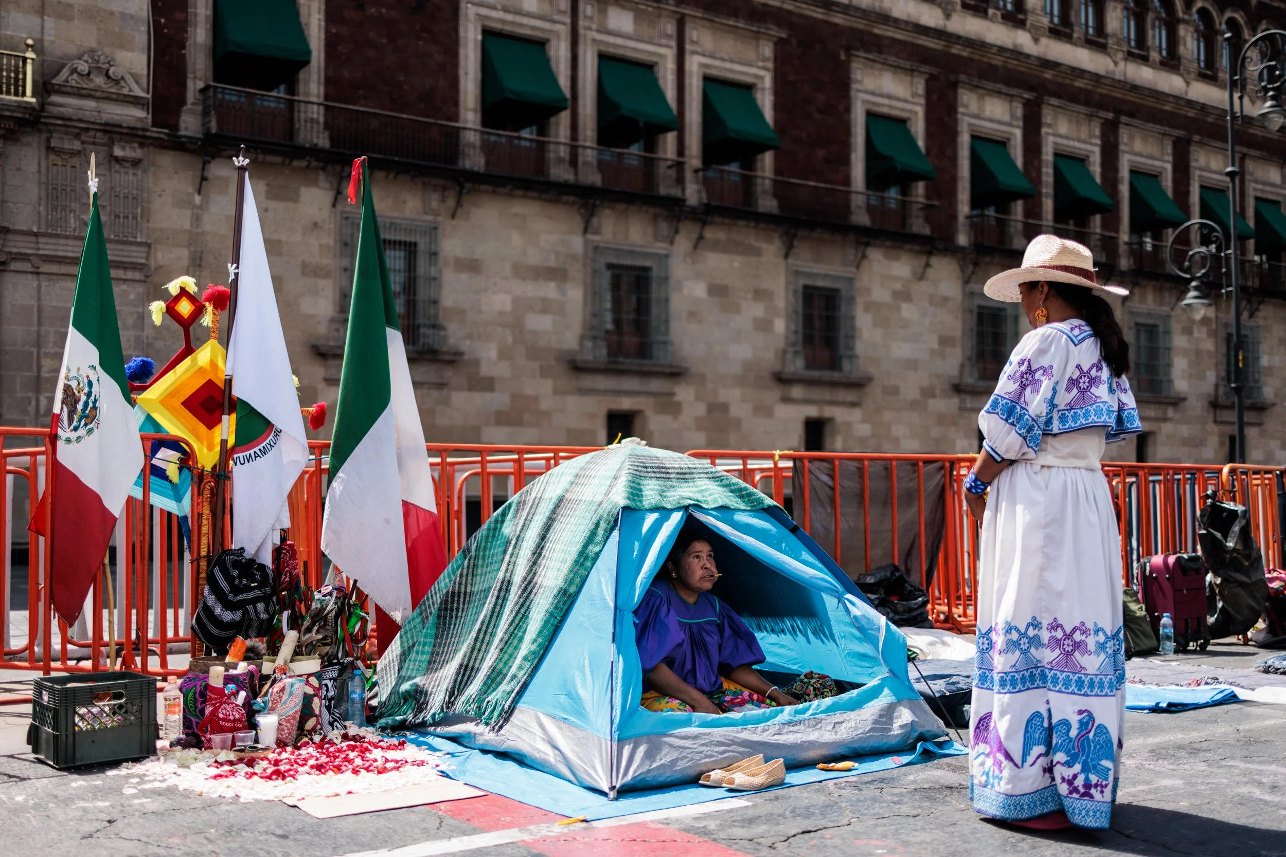 A street scene depicting a woman in traditional Mexican attire standing in front of a homeless woman sitting inside a small tent. The scene includes several Mexican flags and other colorful items for sale or display, with a historic building with gre