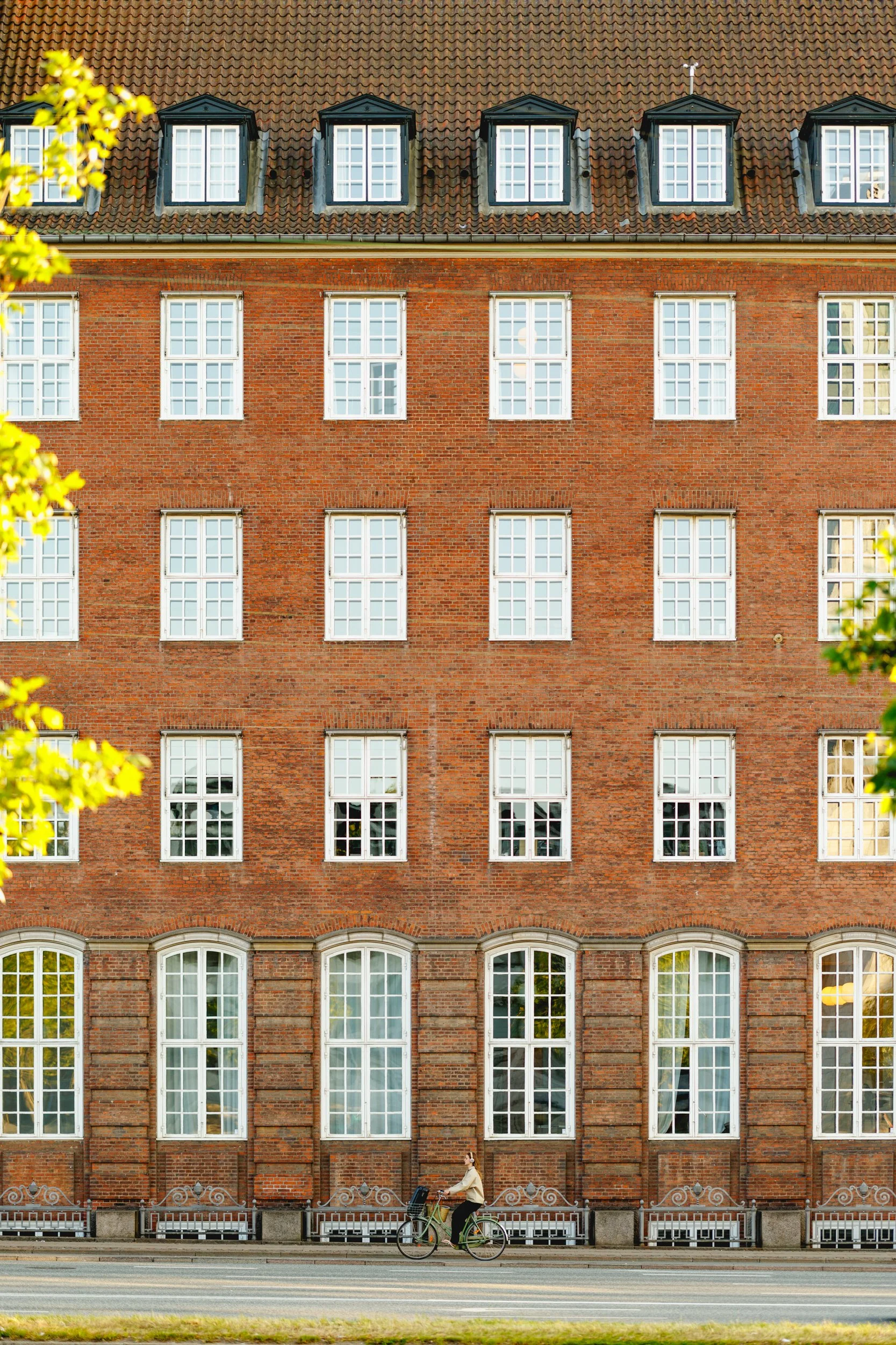 A woman riding a bicycle in front of a red brick multi-story building with large white-framed windows and a tiled roof. Greenery partially visible on the sides.