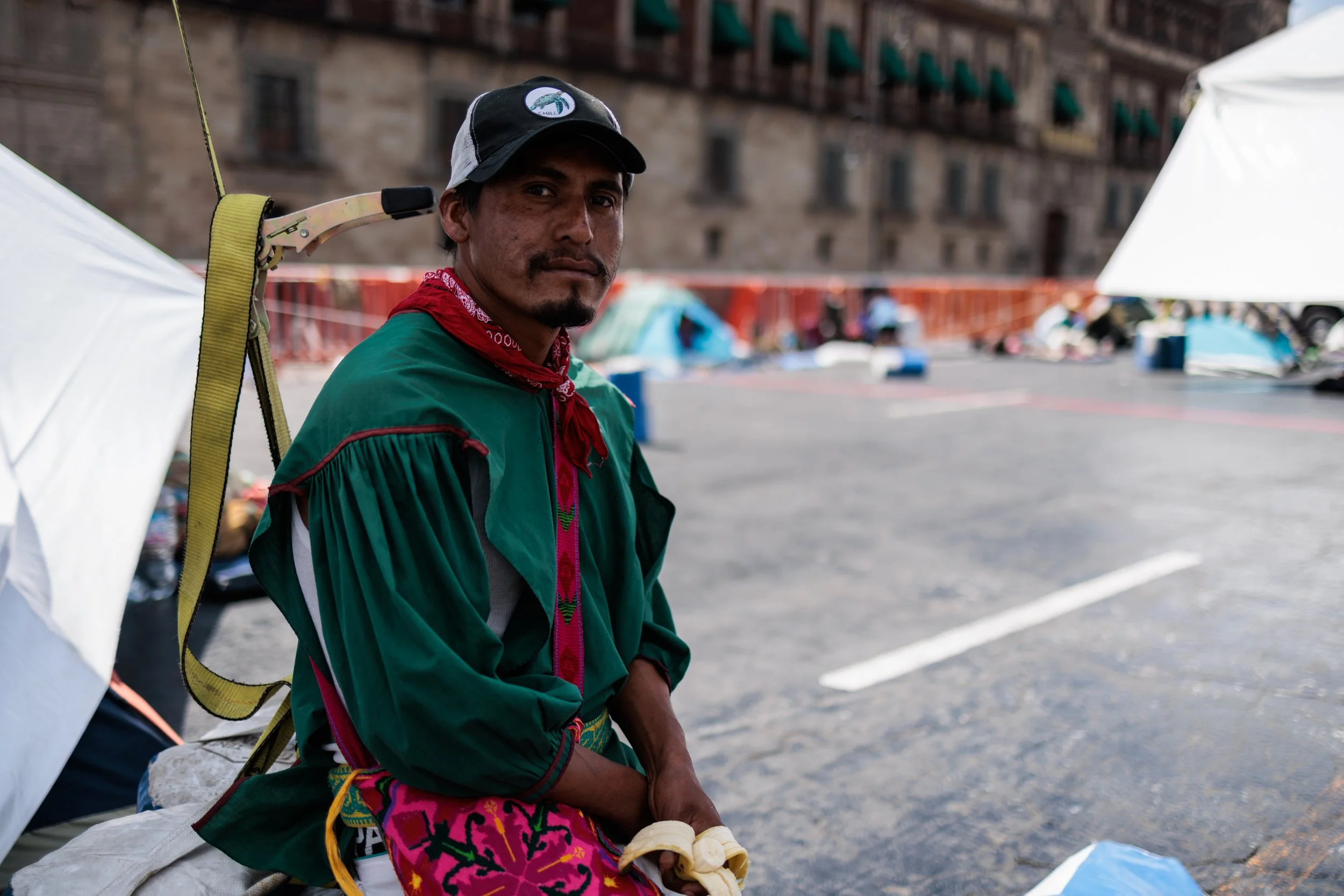 A man wearing a baseball cap, a red bandana, and a green jacket, sitting outdoors near tents and buildings in the background.