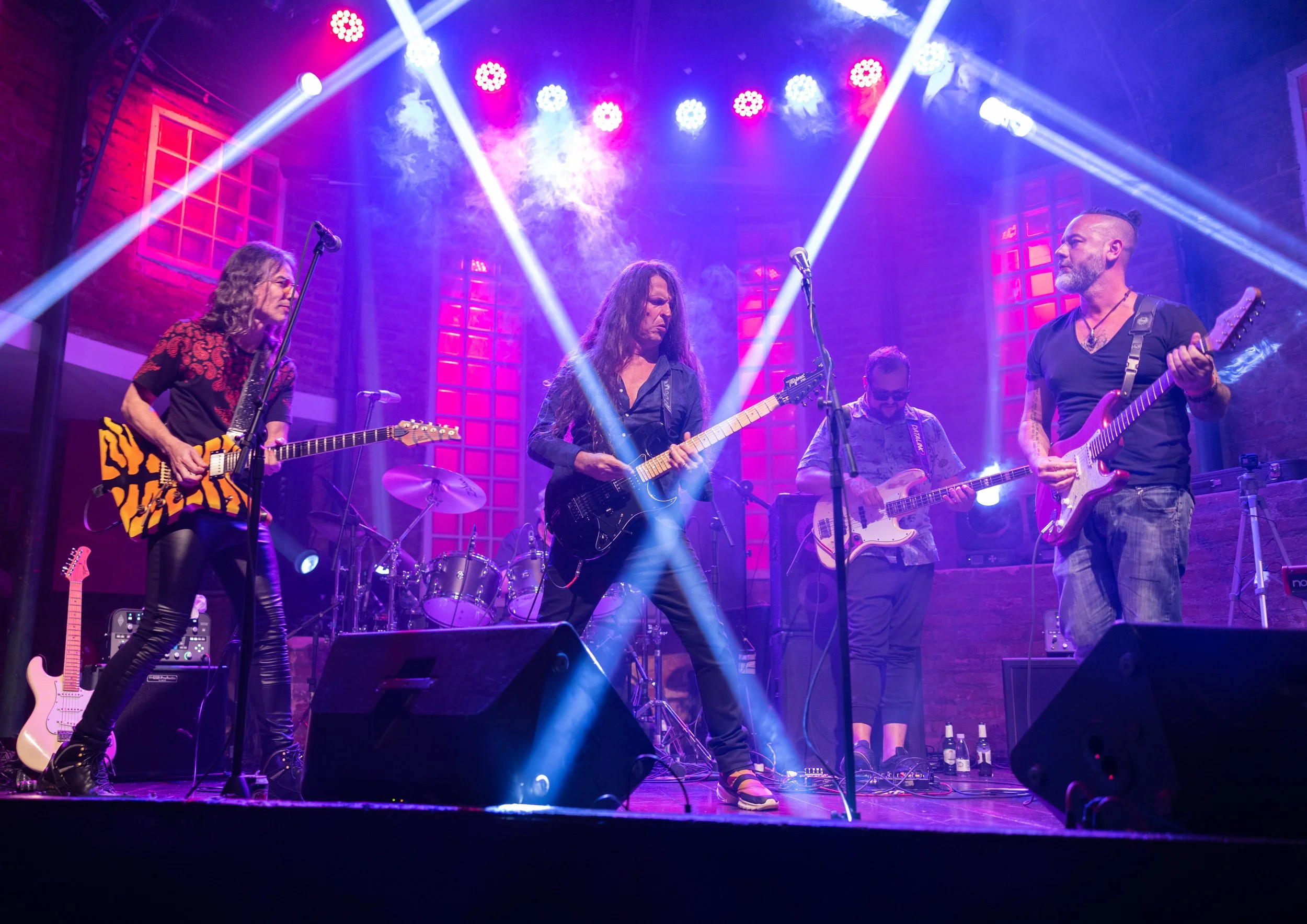 A live band performing on stage with colorful lights and smoke effects, including two guitarists, a keyboardist, and a drummer.