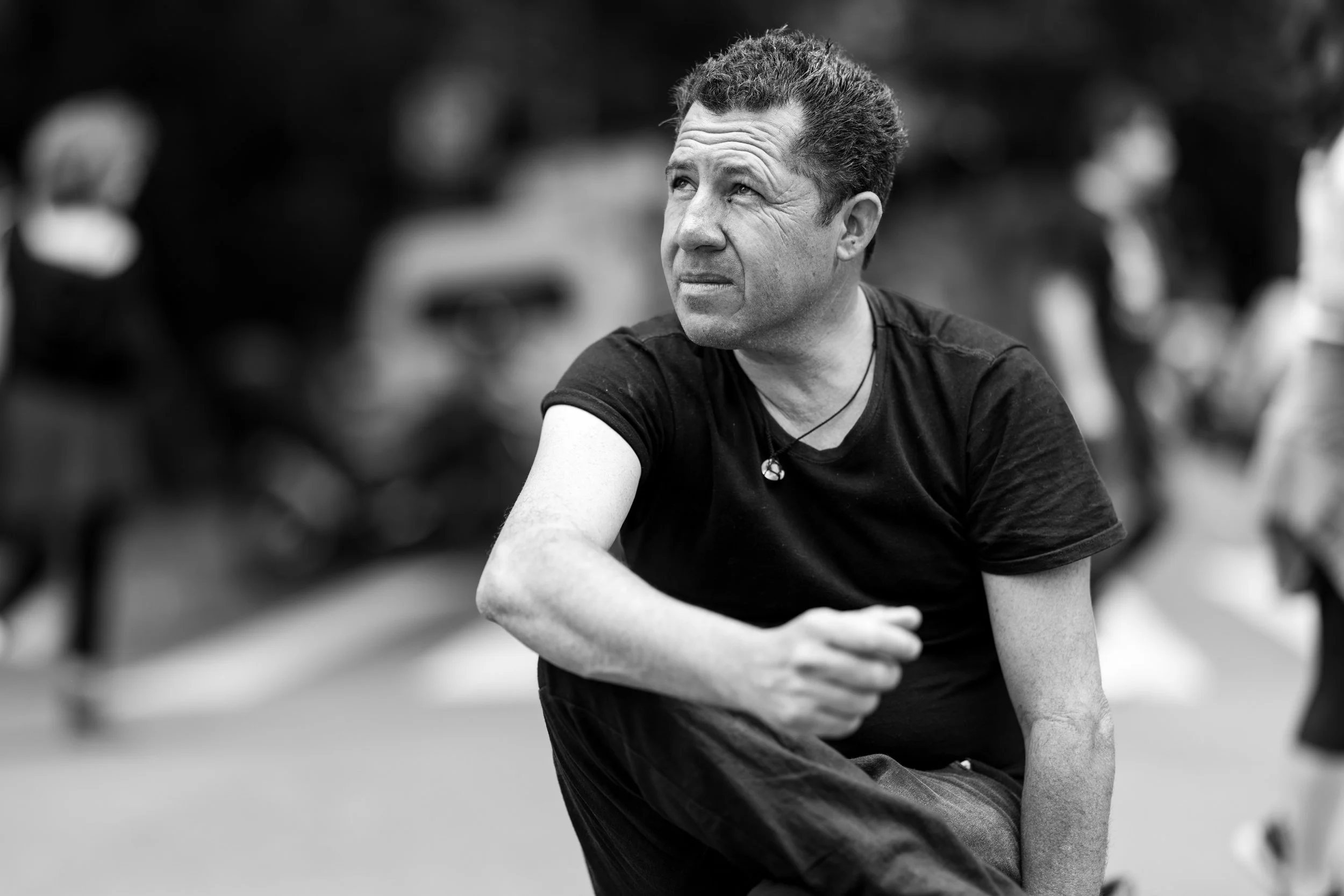 A man with curly hair and a necklace sitting outdoors, looking thoughtfully to the side, with blurred figures in the background.