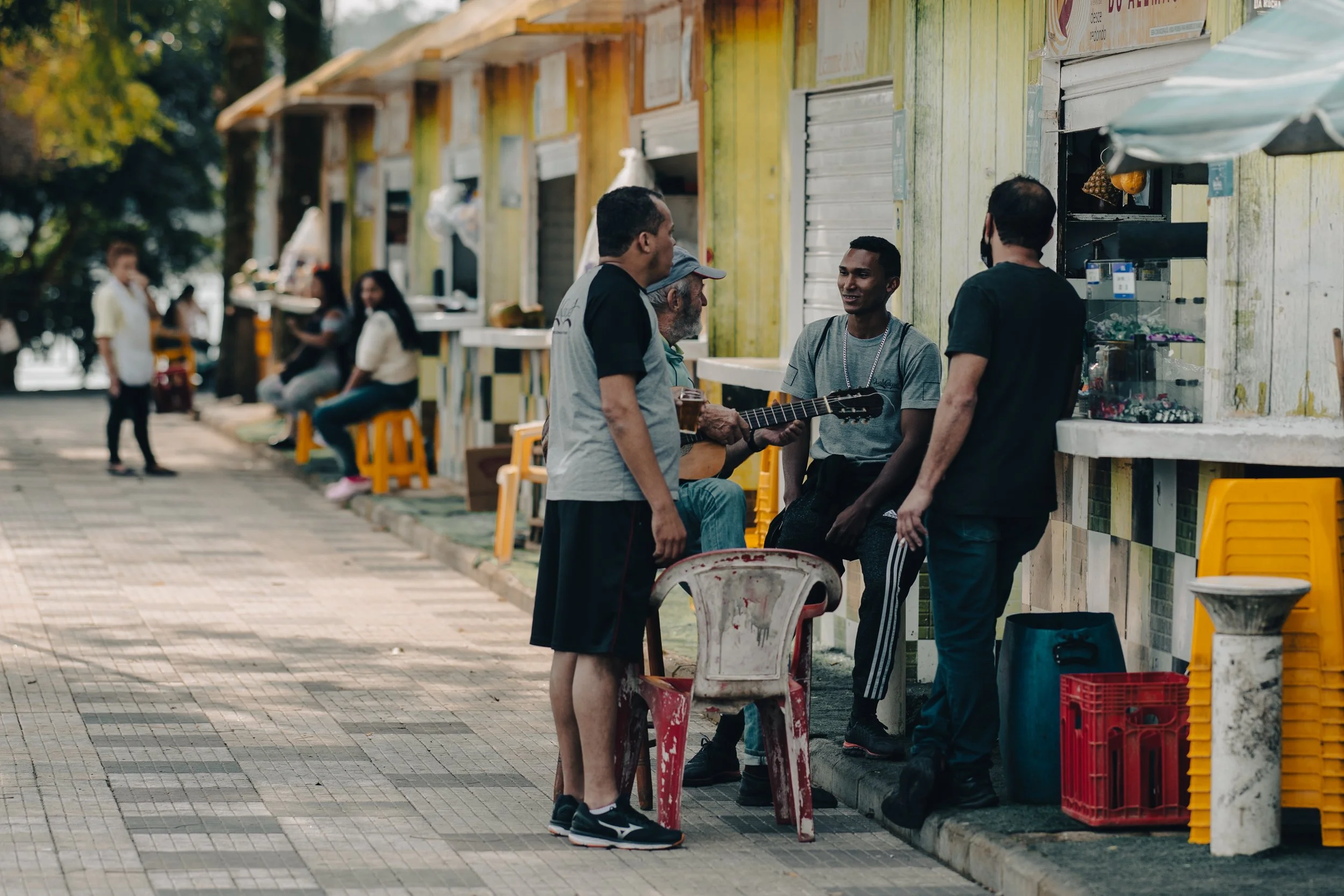 Group of people socializing outside a small food stall with yellow and green walls, some people sitting on yellow stools, and others standing and engaging in conversation.