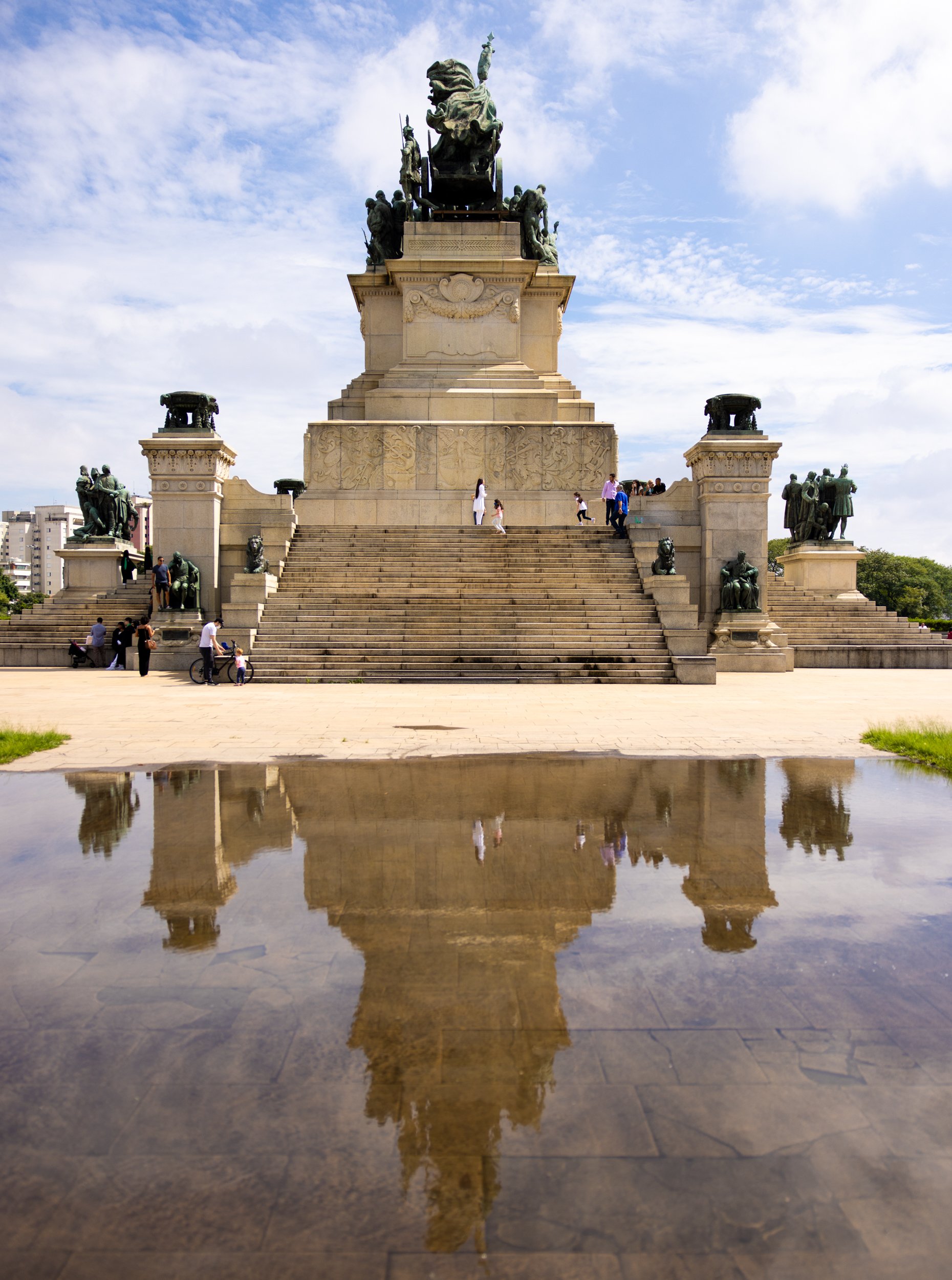 A large monument with stairs, sculptures, and a central figure on top, against a partly cloudy sky, with people walking and gathering around.