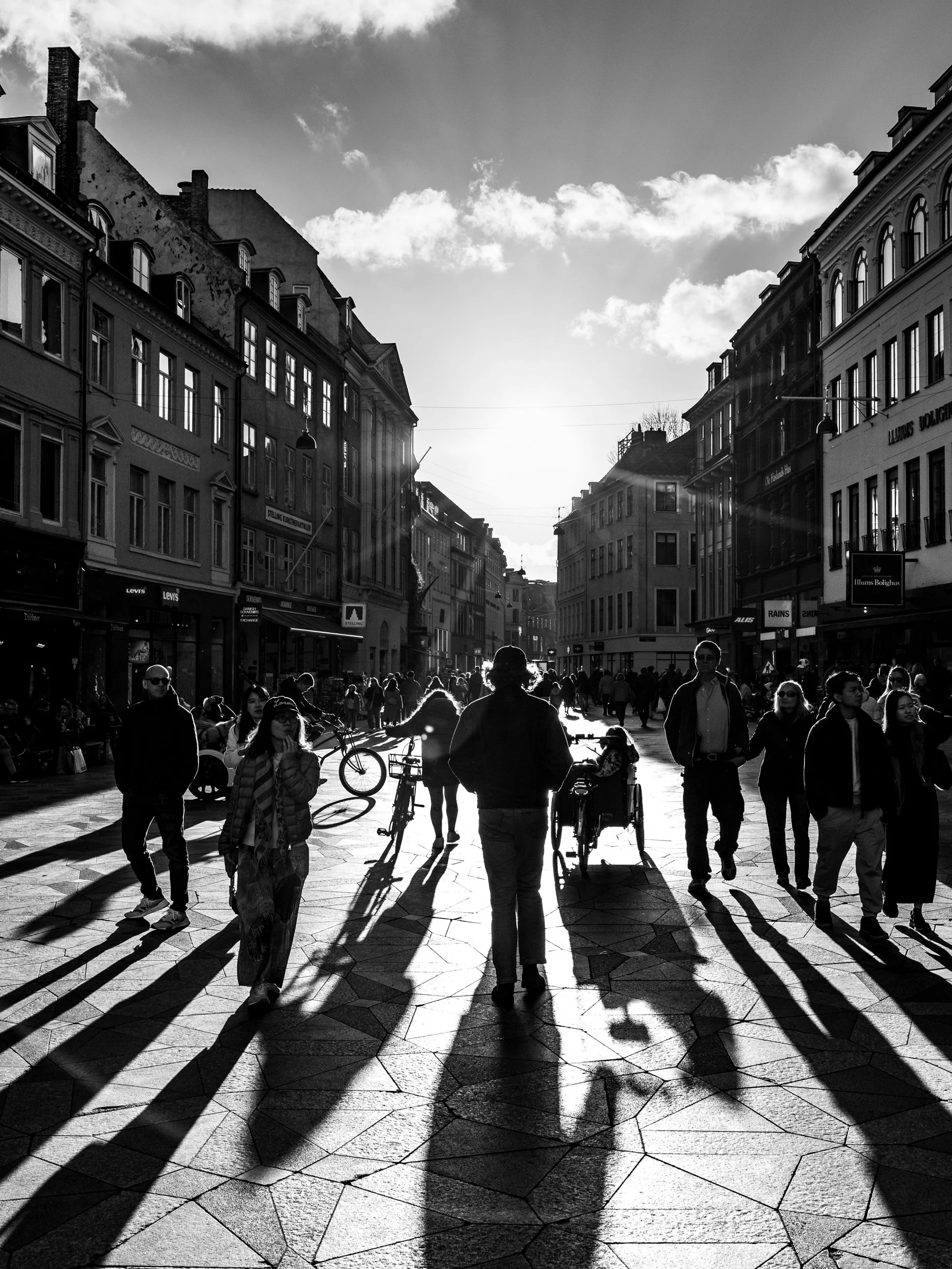 Black and white photo of a city street with buildings on either side, people walking and riding bikes, and long shadows on the ground from the low sun in the sky.