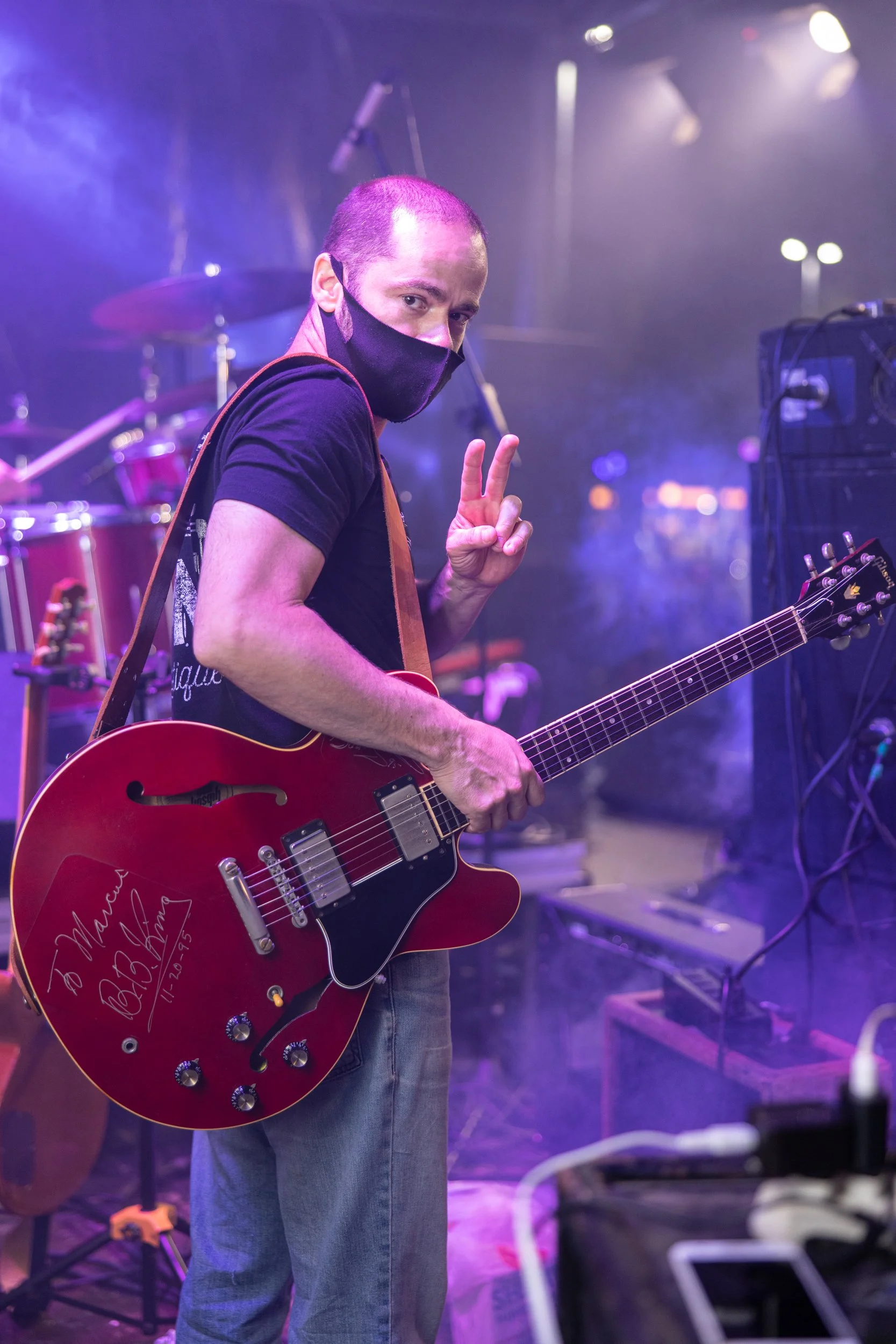A man with a shaved head and wearing a black mask is giving a peace sign while holding a red guitar with autographed inscriptions. He is on stage with drum equipment and stage lighting visible in the background.