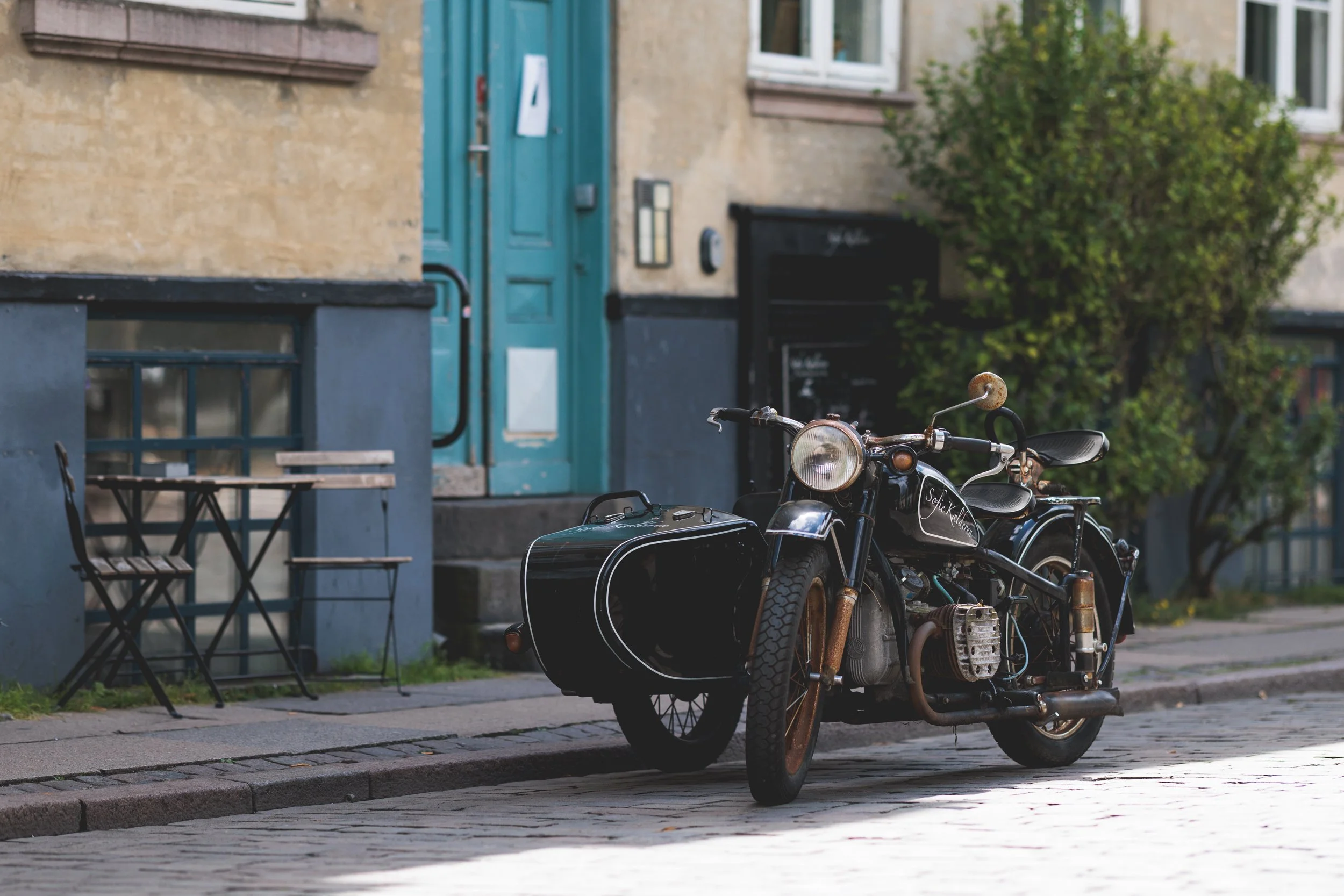 A vintage black motorcycle with sidecar parked on a cobblestone street in front of a building with a green door and windows. There are outdoor tables and chairs nearby, and a bush is visible in the background.