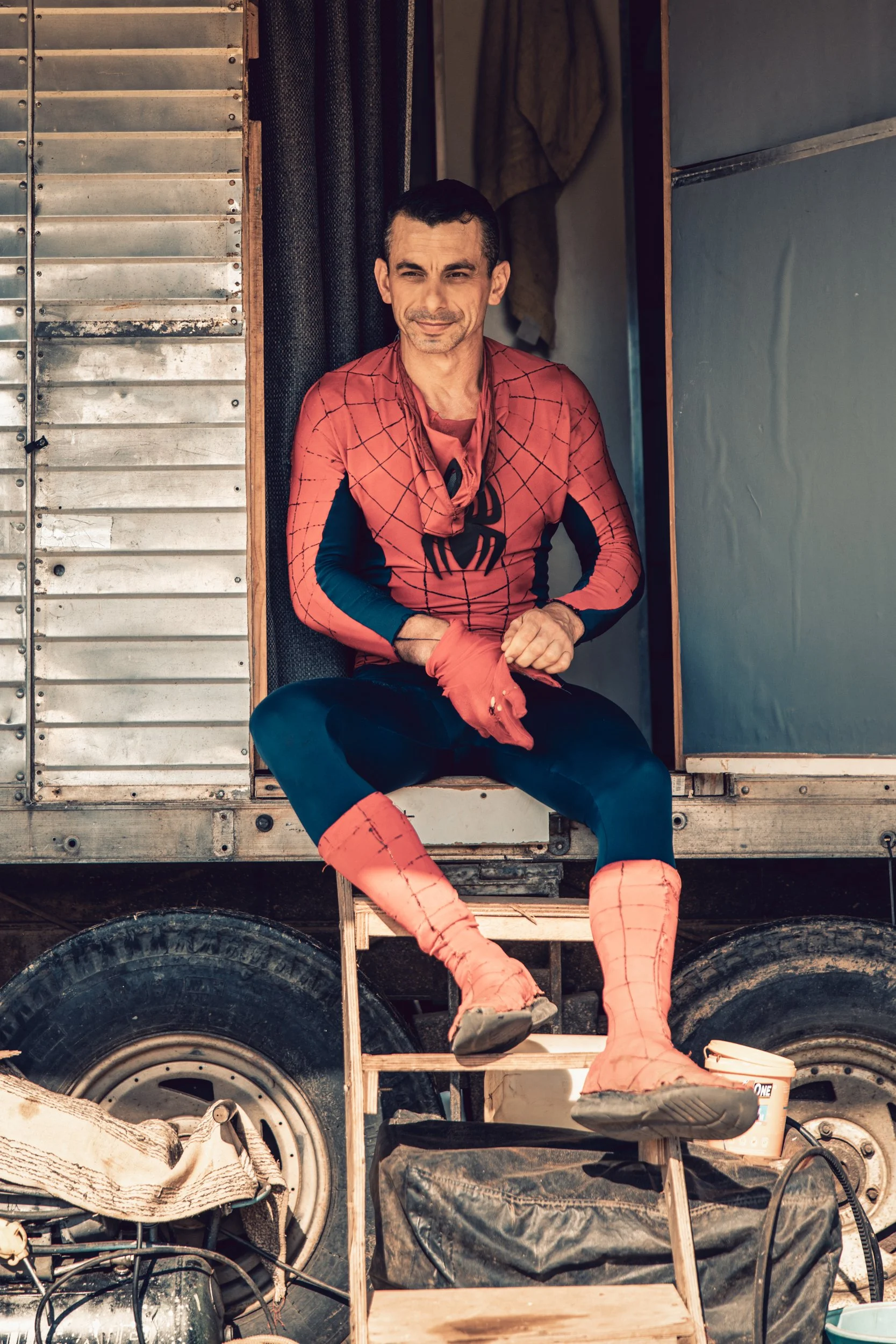 A man dressed as Spider-Man sitting on a wooden step in front of a wooden structure, surrounded by tires and various objects.