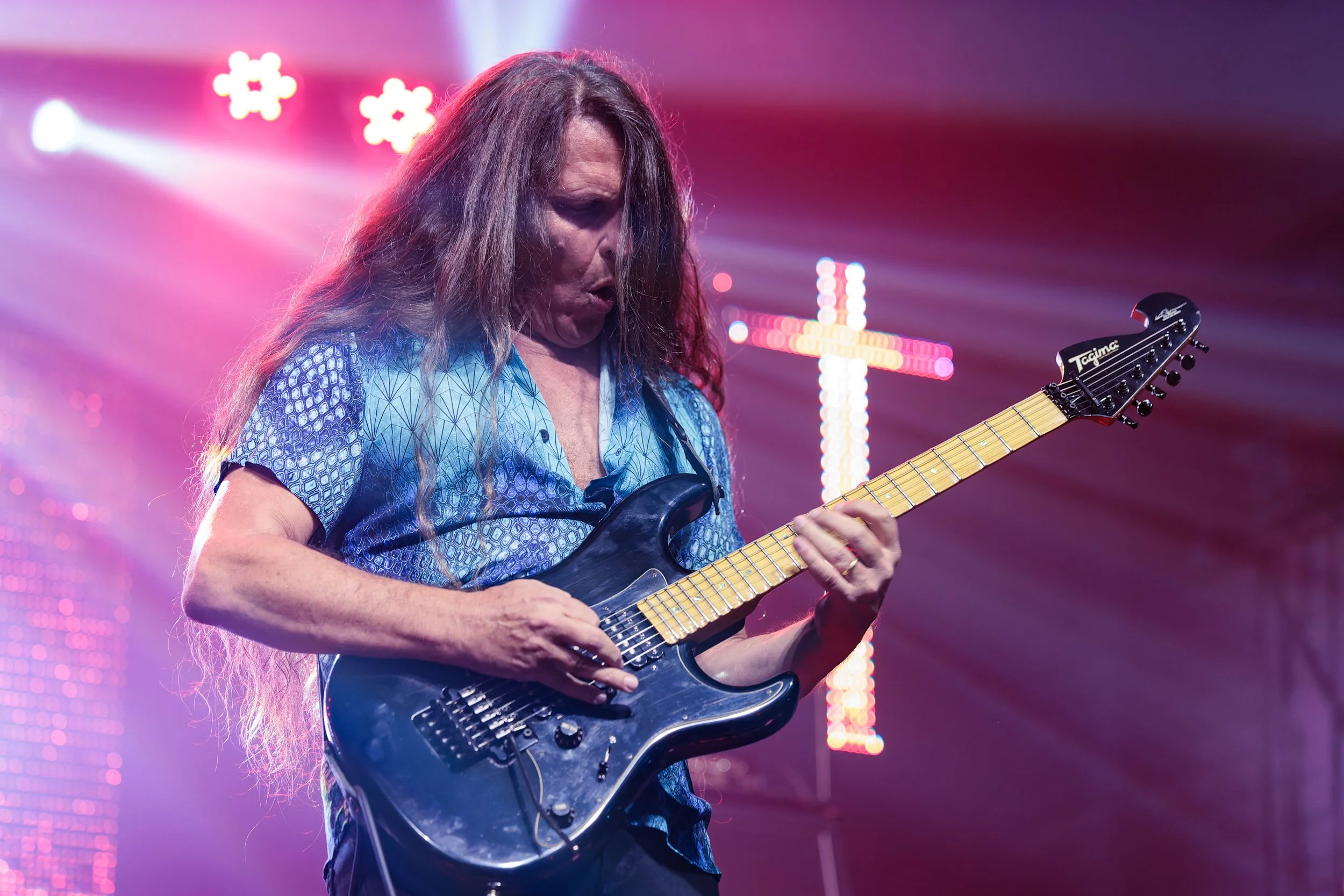 A woman with long hair playing an electric guitar on stage with colorful lights in the background.