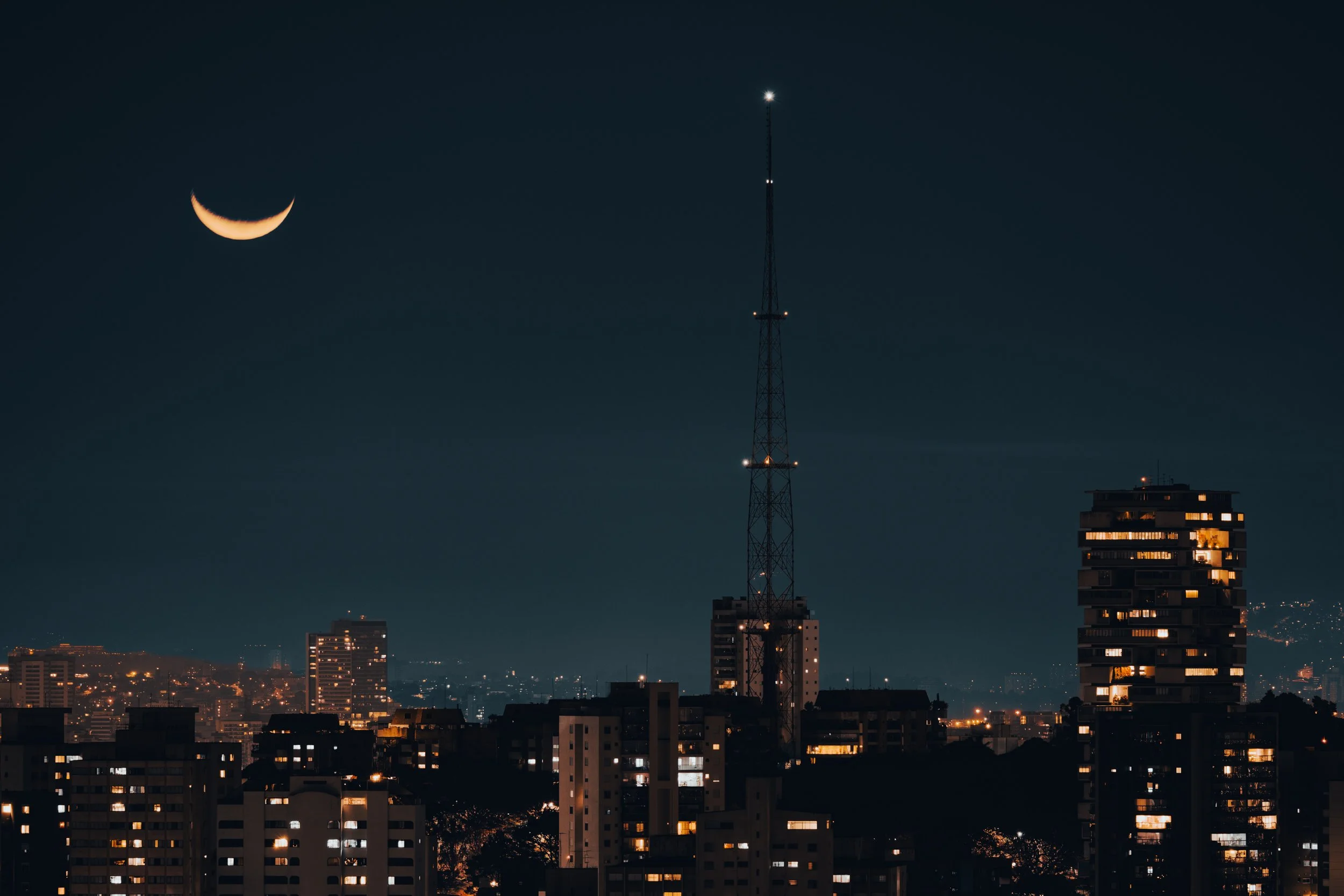 Nighttime cityscape featuring a tall radio tower with lights and a crescent moon in the sky, buildings with lit windows below.