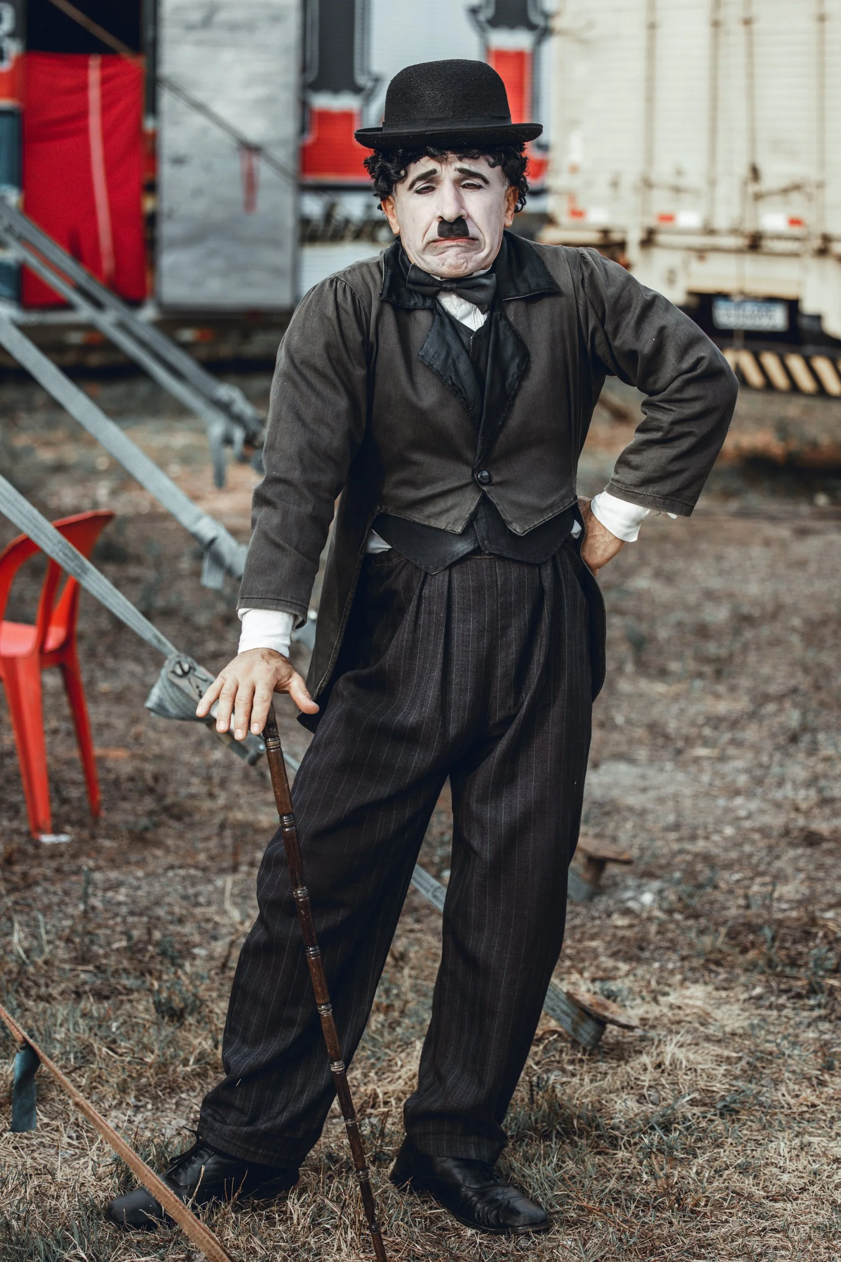 A man dressed as Charlie Chaplin with a sad facial expression, wearing a black bowler hat, a black jacket, and striped trousers, standing outdoors in front of train cars and equipment.