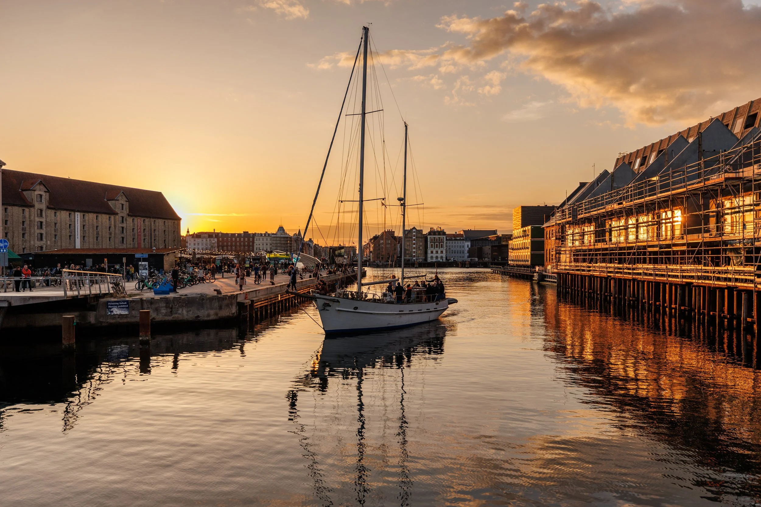 A scenic harbor at sunset with boats docked, people walking along the pier, historic buildings, and orange-yellow light reflecting on the water.