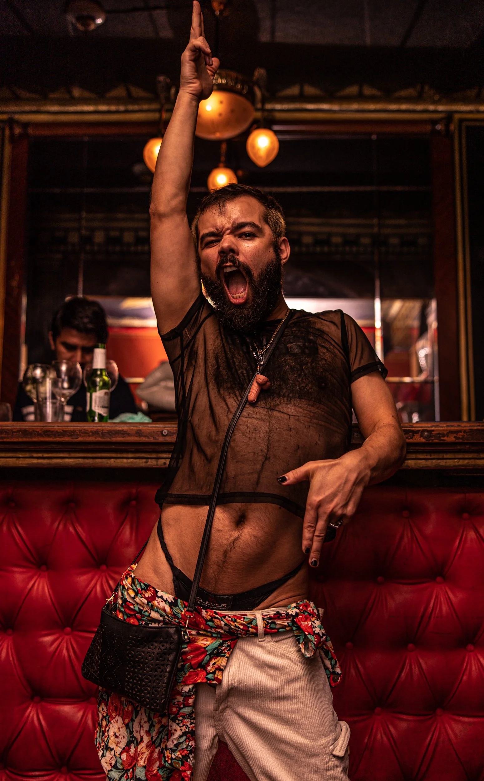 A man posed provocatively in a bar, wearing a see-through black mesh shirt, floral shorts, and beige cargo pants, with a black crossbody bag, raising one arm and shouting.