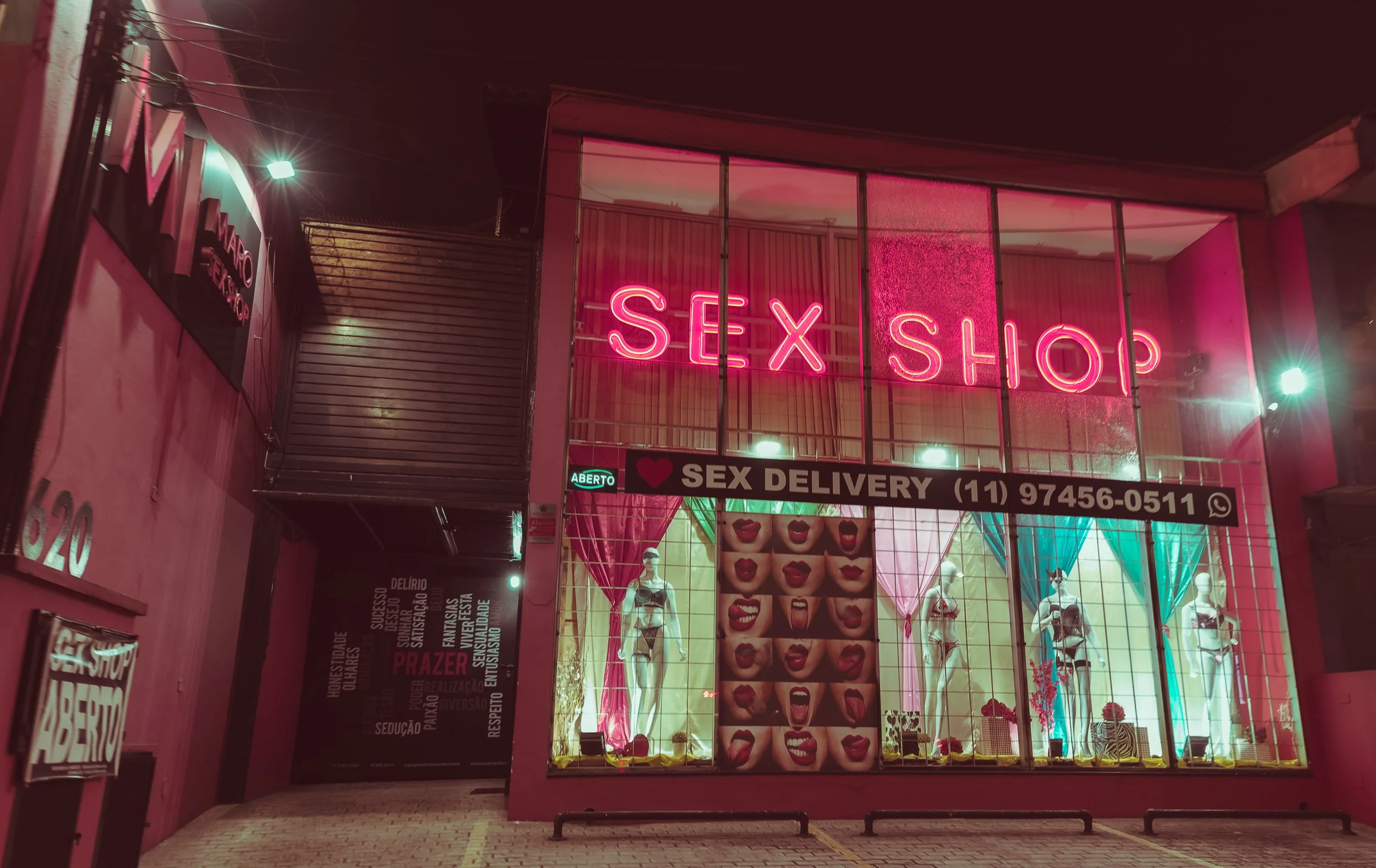 Storefront with a pink neon sign that says 'SEX SHOP' at night, large window display with mannequins and art of mouths with various lipstick colors, and signs indicating 'SEX DELIVERY' and an open sign.