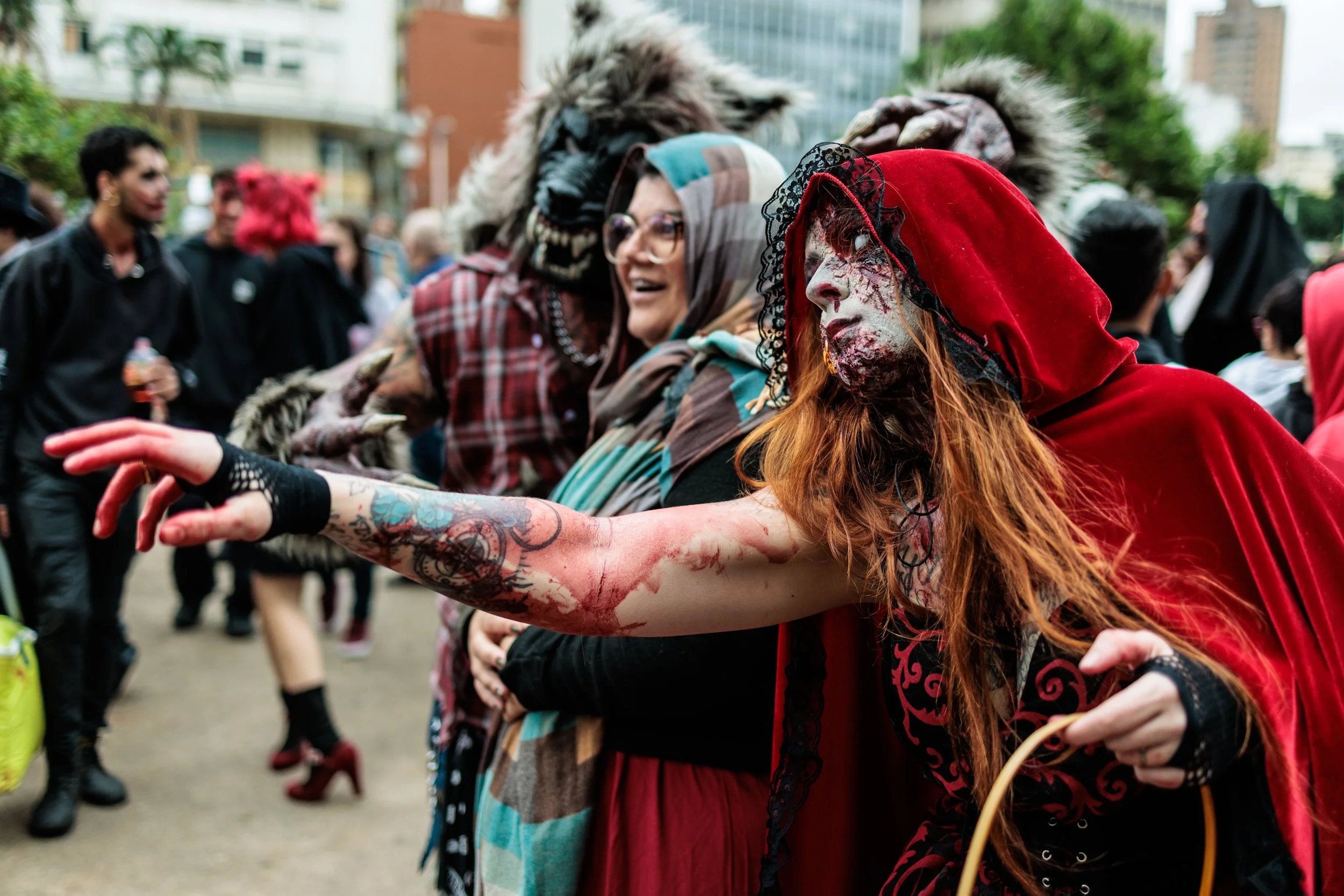 People in costumes, including a woman with a zombie makeup and a red hood, participate in a street event or festival with an urban backdrop.