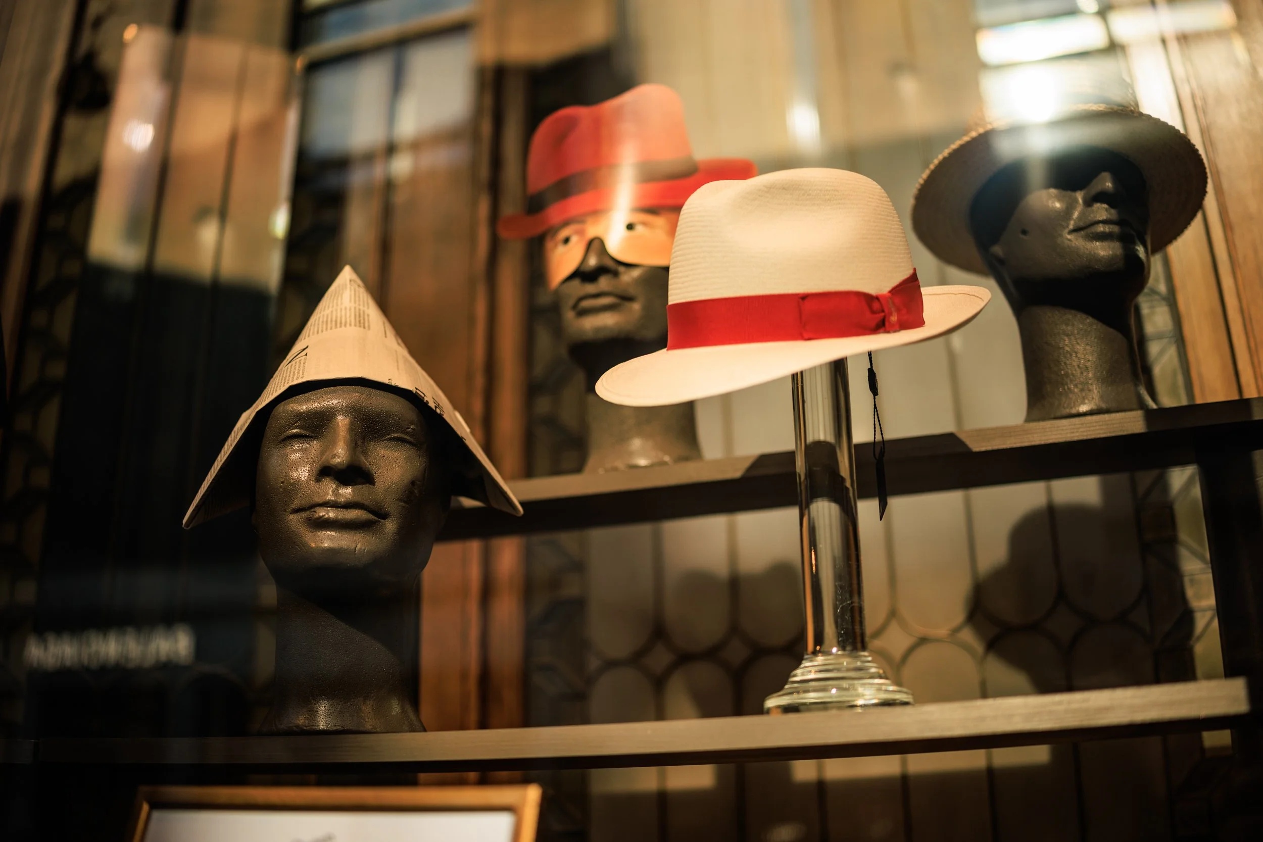 Display of mannequin heads and hats, including a white hat with a red ribbon, a black hat with wide brim, and a red hat with black band, on shelves with wooden and glass backgrounds.