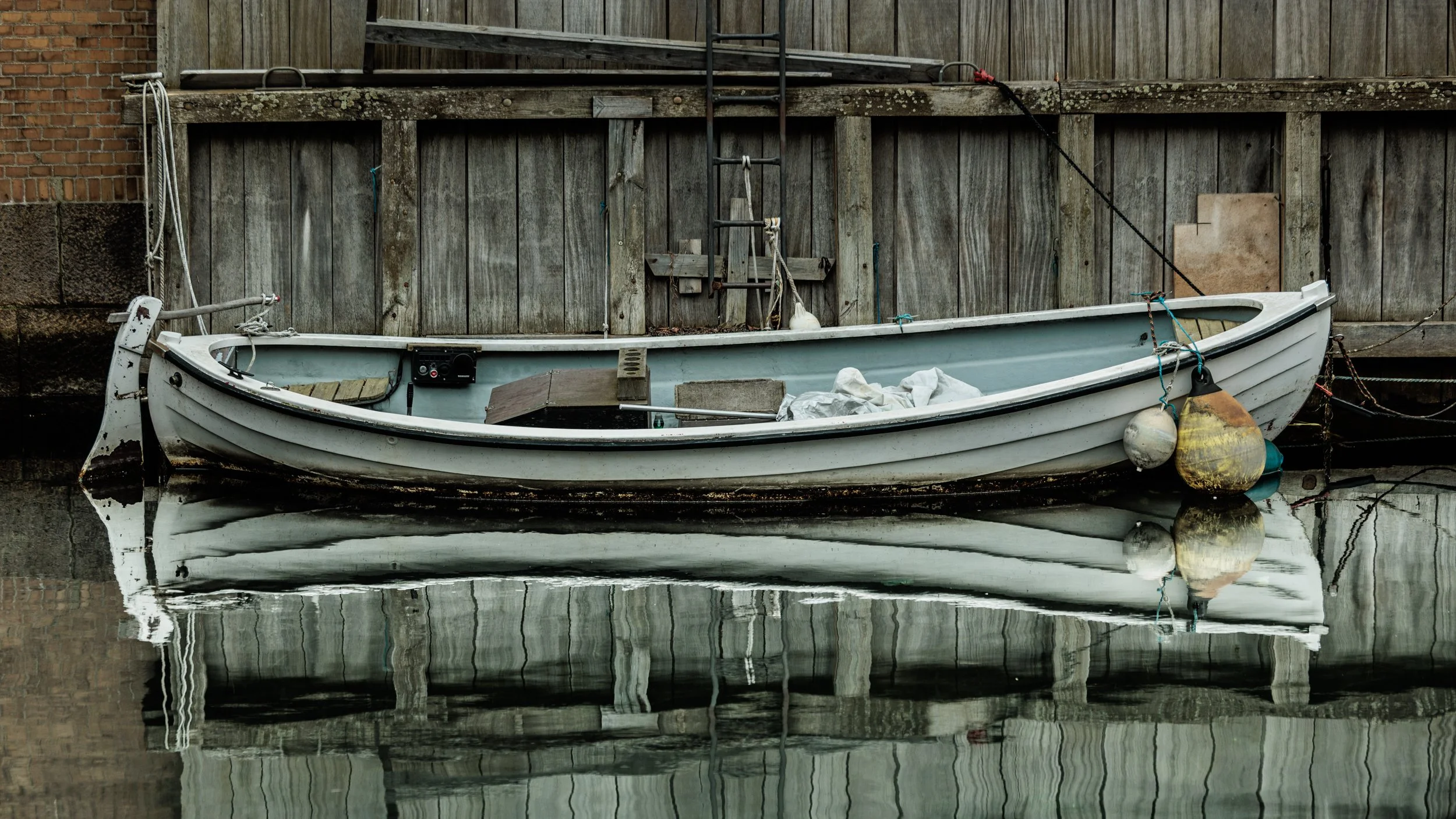 An old white rowboat moored beside a weathered wooden dock with fishing buoys hanging from its side, reflected in calm water.