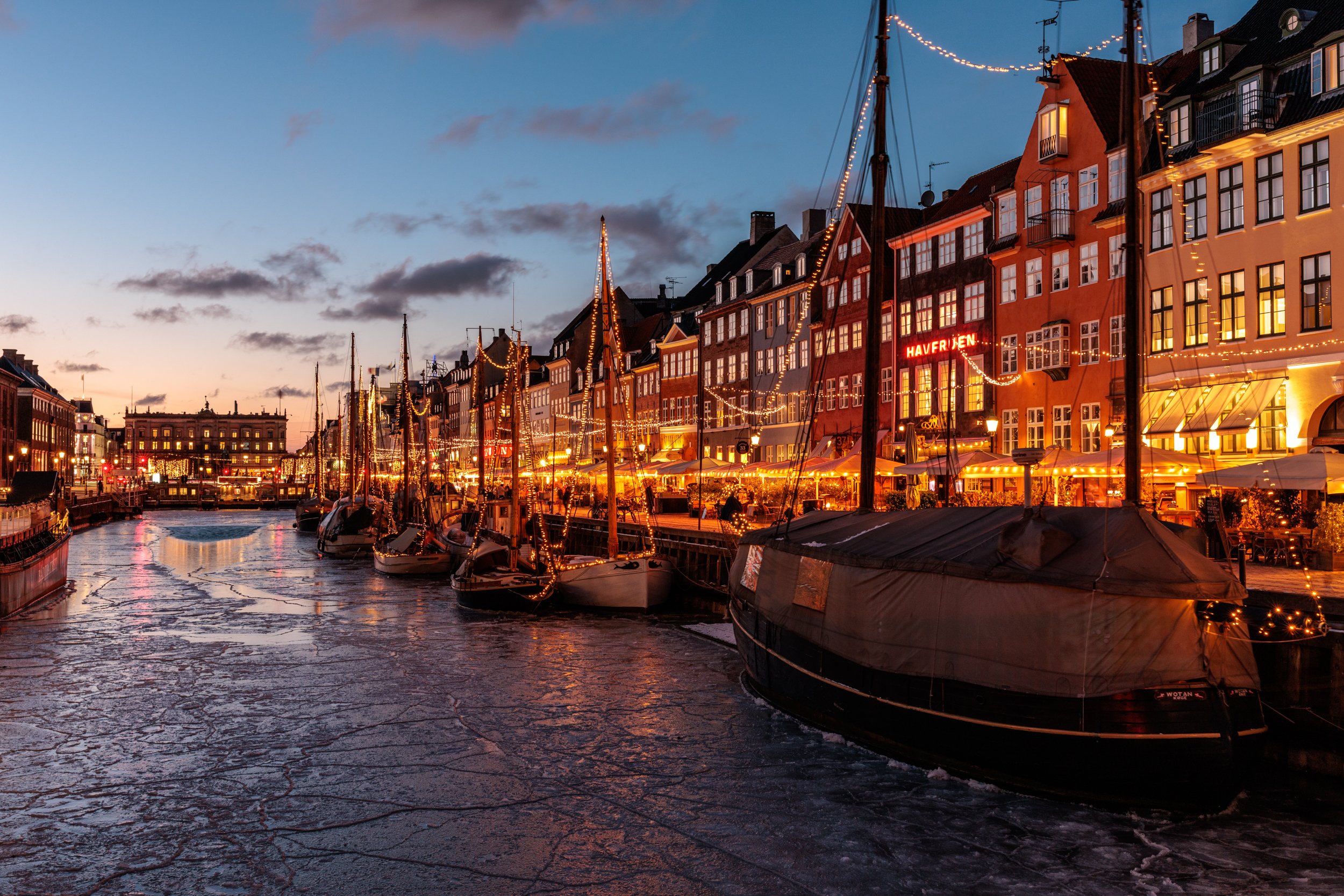 A harbor at dusk with boats docked along the pier, buildings decorated with string lights, and a sky transitioning from sunset to night.