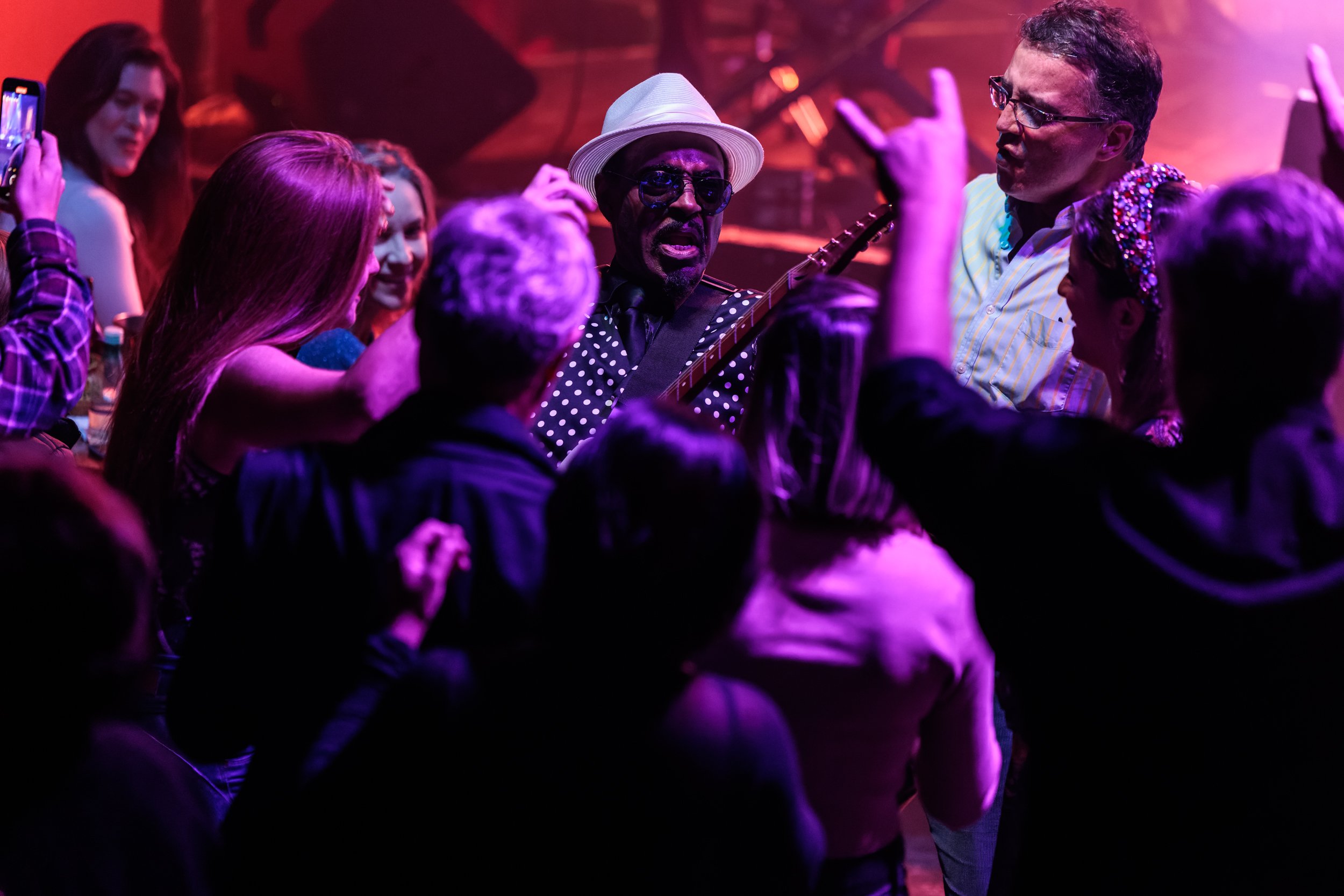 A group of people dancing and socializing at a party or nightclub with purple and pink lighting. A man in sunglasses and a white hat is at the center, playing a guitar, while others around him are smiling and enjoying the moment.