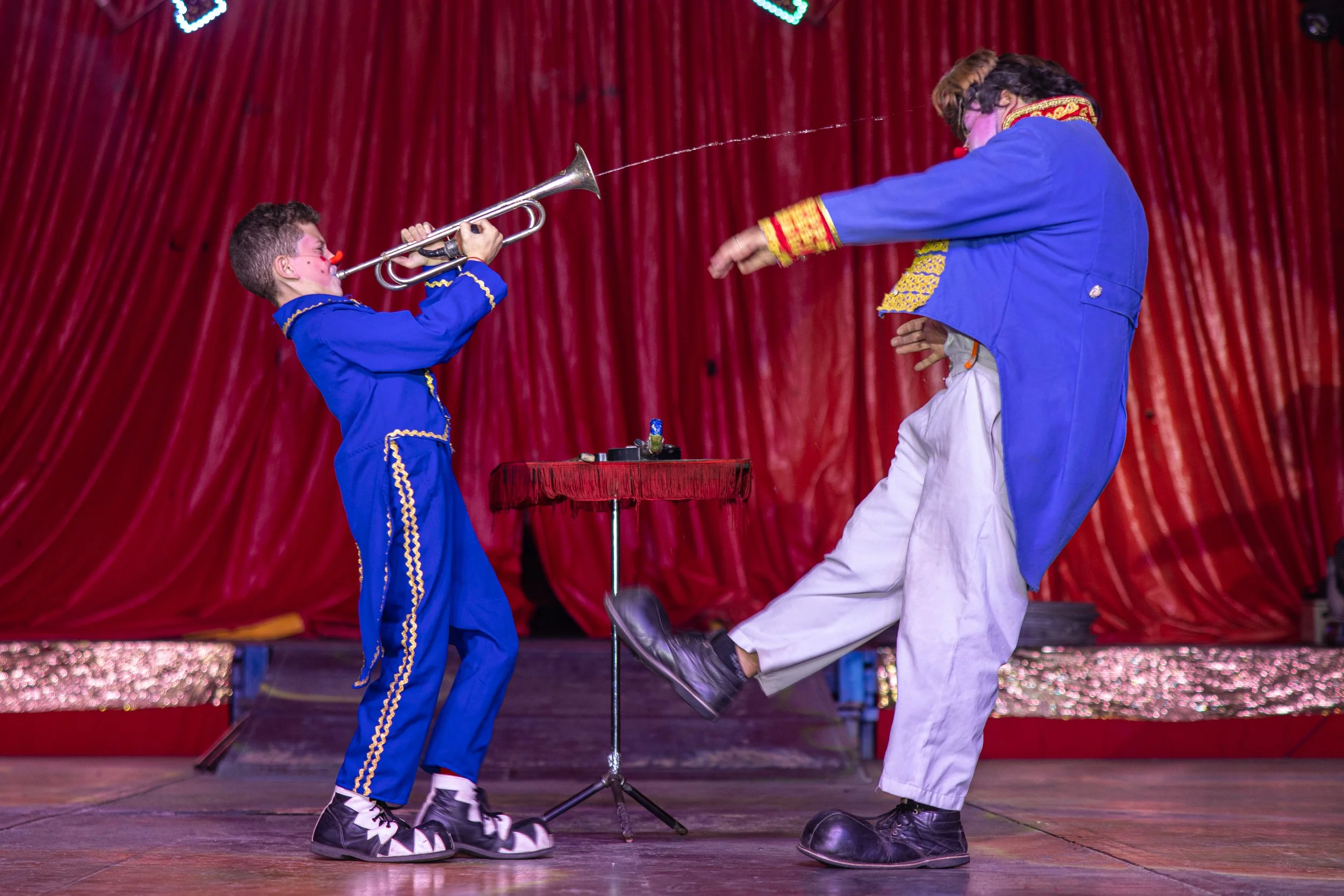 A clown and a young boy in blue costumes perform a routine on stage with a red curtain backdrop. The boy is blowing into a horn, and the clown is holding a wand with a string attached to the horn, with various props on a small table in the background