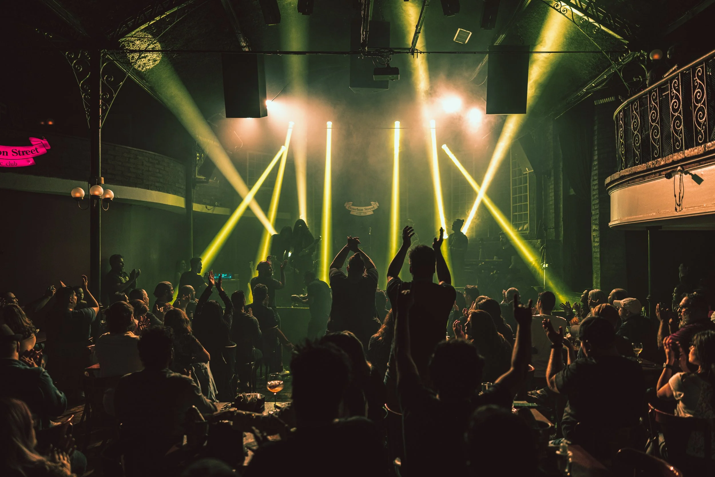 A concert or nightclub scene featuring a crowd of people dancing and enjoying a live music performance. Colorful stage lights, including yellow beams, illuminate the dark venue. The stage has large speakers and a band performing, with a pink sign on the side that says 'Bourbon Street.'