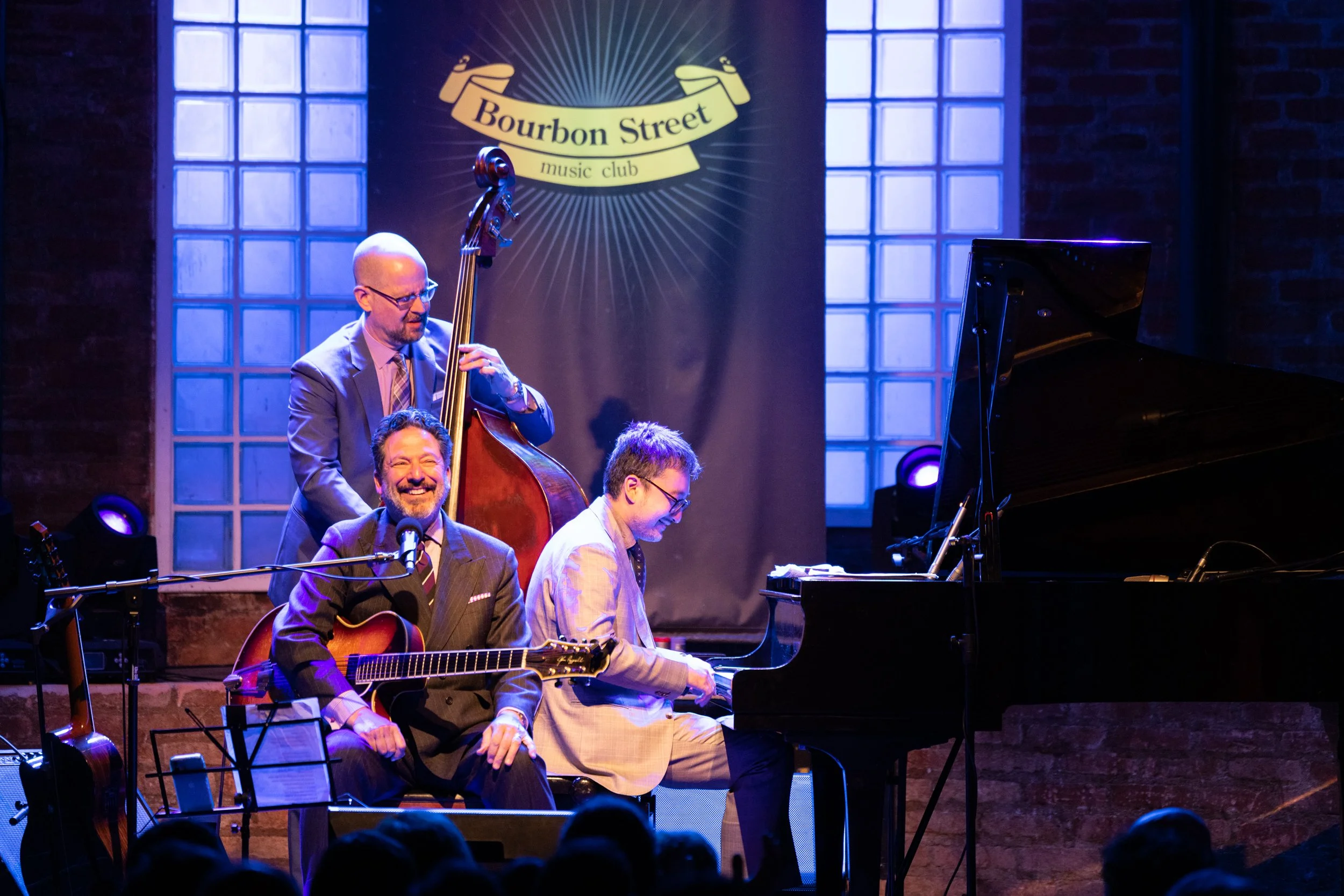 Jazz band performing at Bourbon Street music club with four musicians, including a guitarist, a pianist, a double bassist, and a saxophonist, under blue and purple stage lighting.