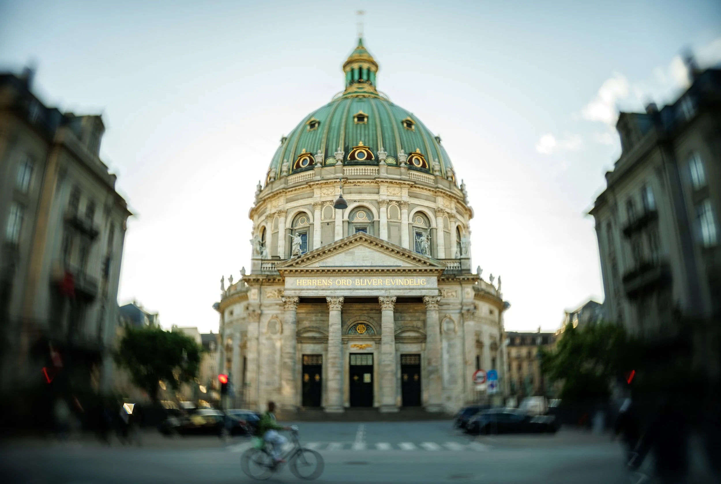 A large historic building with a green dome topped with a golden spire, flanked by ornate statues and columns, captured from a street view with blurred cars and a cyclist in motion.